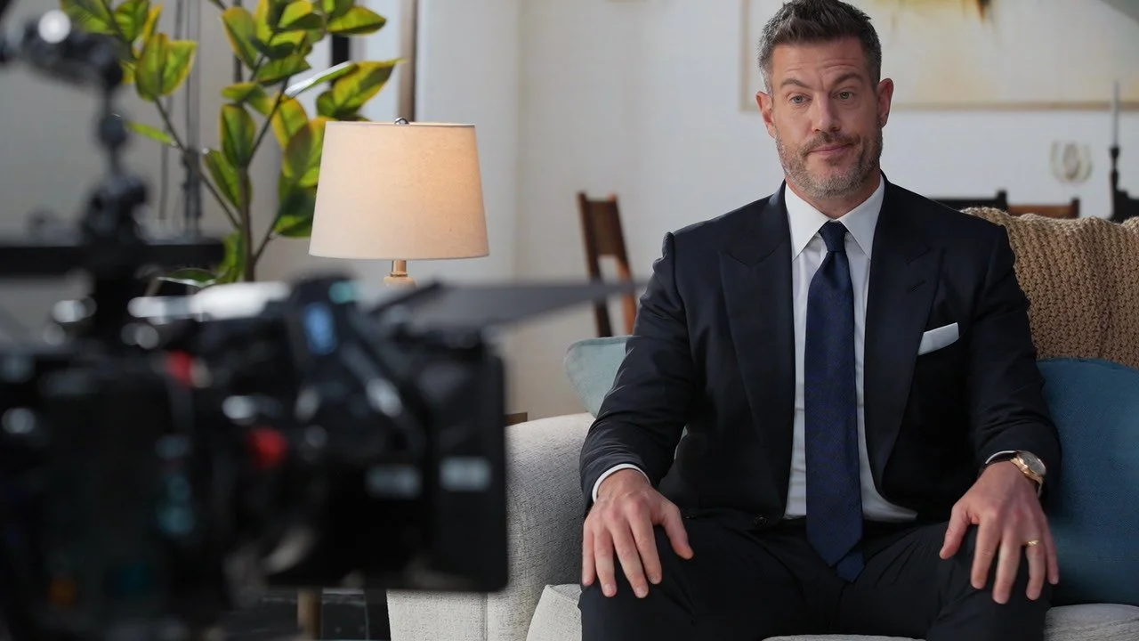 A man in a black suit and blue tie sitting on a couch in a living room during a video recording or interview, with a camera and recording equipment in front of him.