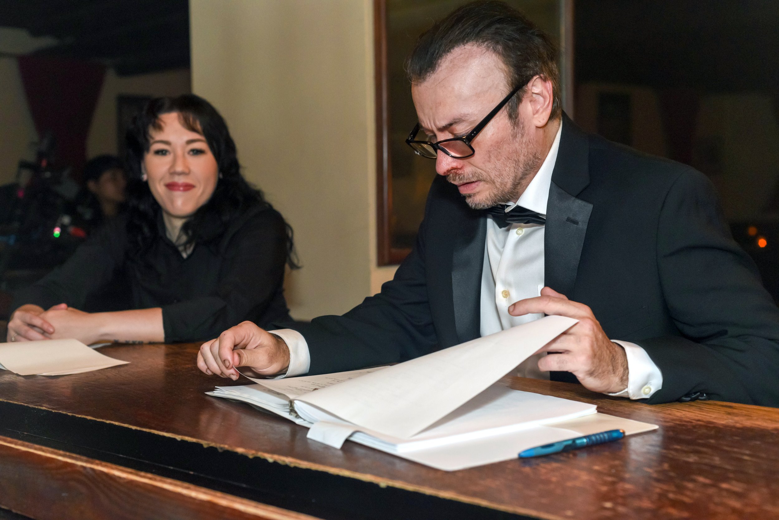 A man in a tuxedo with glasses is looking down and signing papers at a wooden table, with a woman smiling in the background.