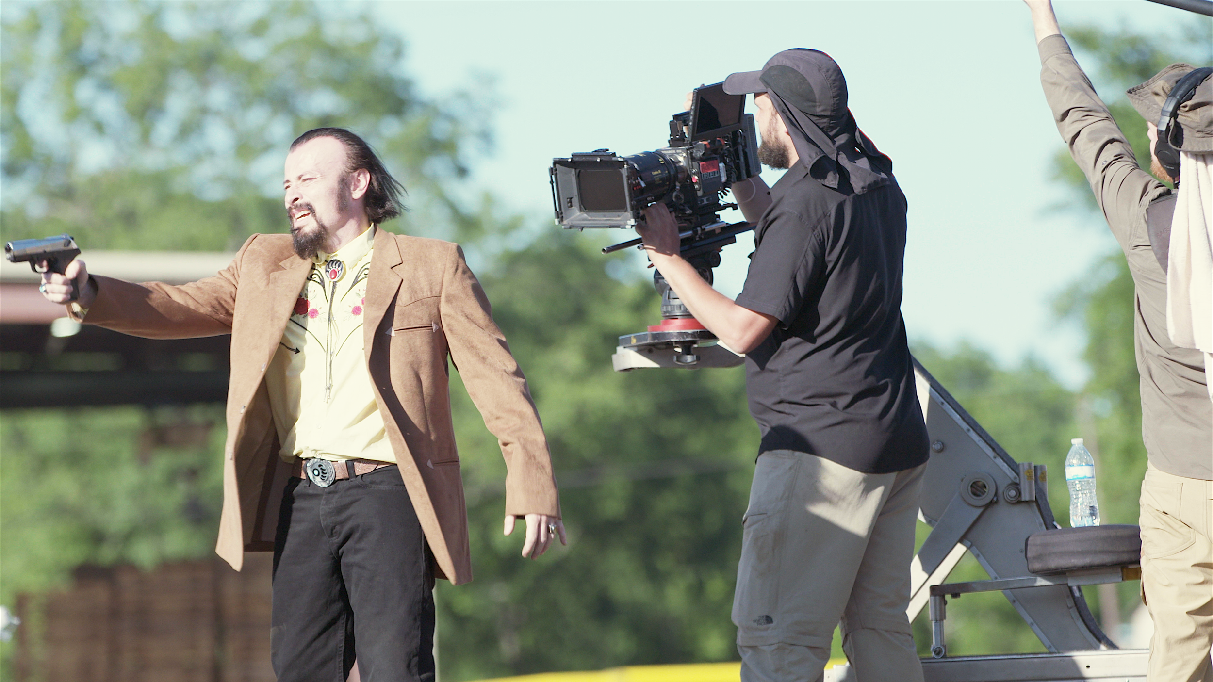 Man holding a gun on a film set with a camera operator filming him outdoors.