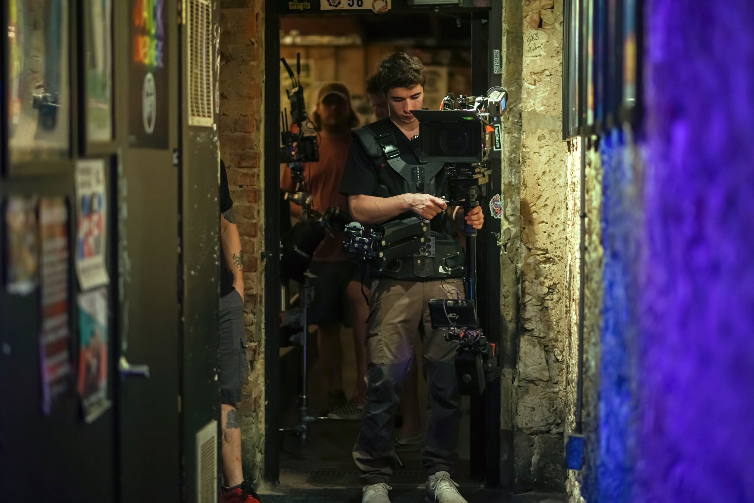 A person standing in front of a professional camera, focused on filming, in a dimly lit area with brick walls and vending machines in the foreground.