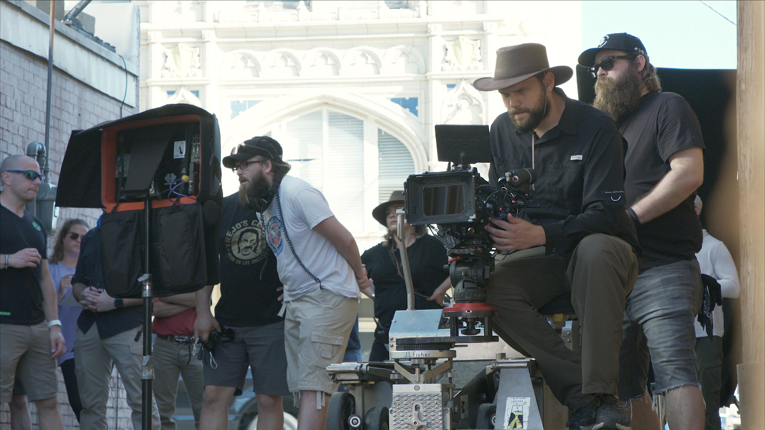 A film crew on a city street, including a man operating a camera on a tripod, another man in a wide-brimmed hat and sunglasses, and other crew members and onlookers nearby.