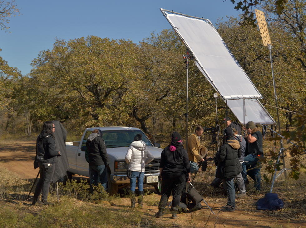 Film crew shooting outdoors in a rural area with trees, using a large reflector and camera equipment in front of a parked pickup truck.