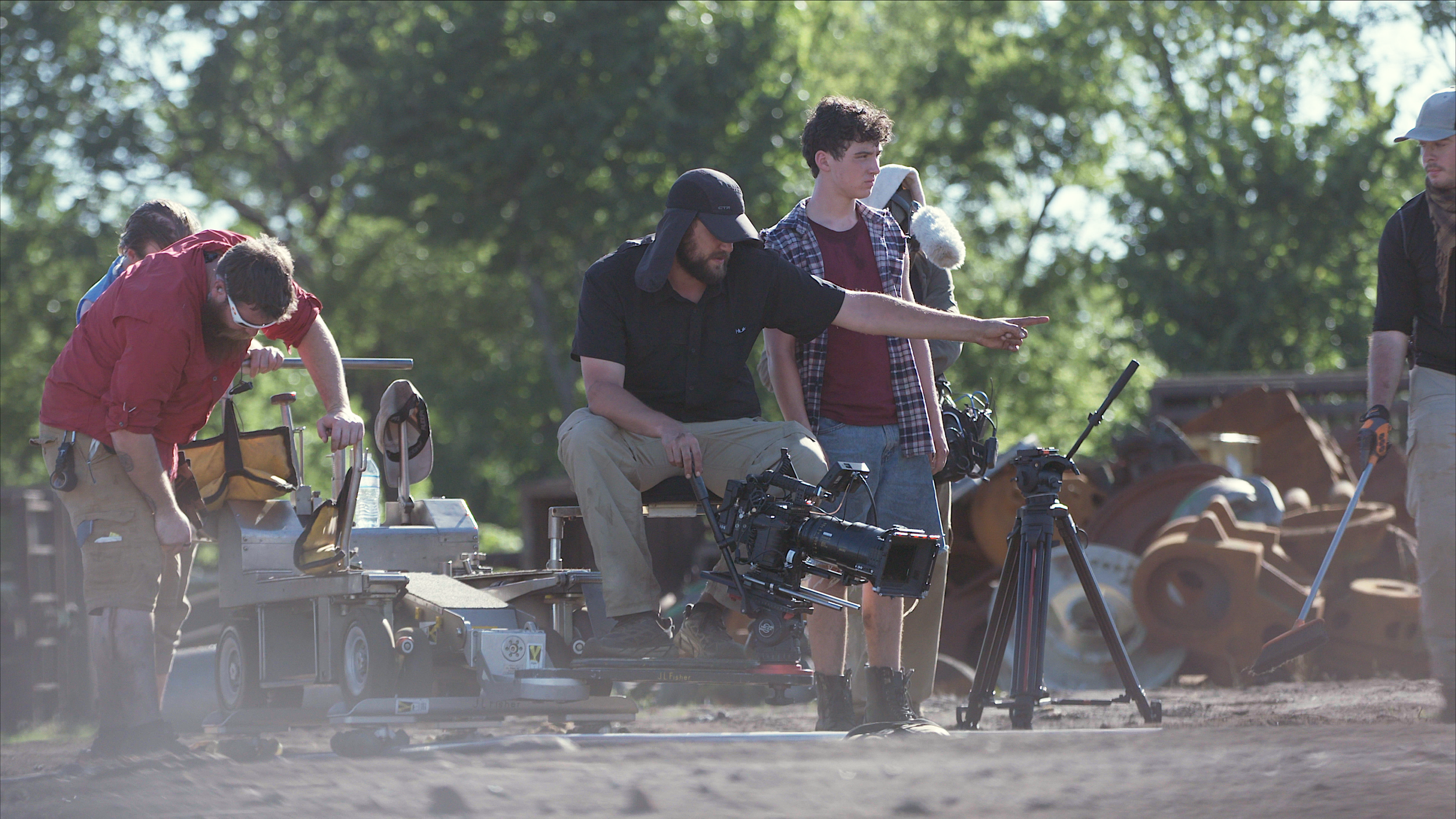 Film crew setting up equipment outdoors with trees in the background.