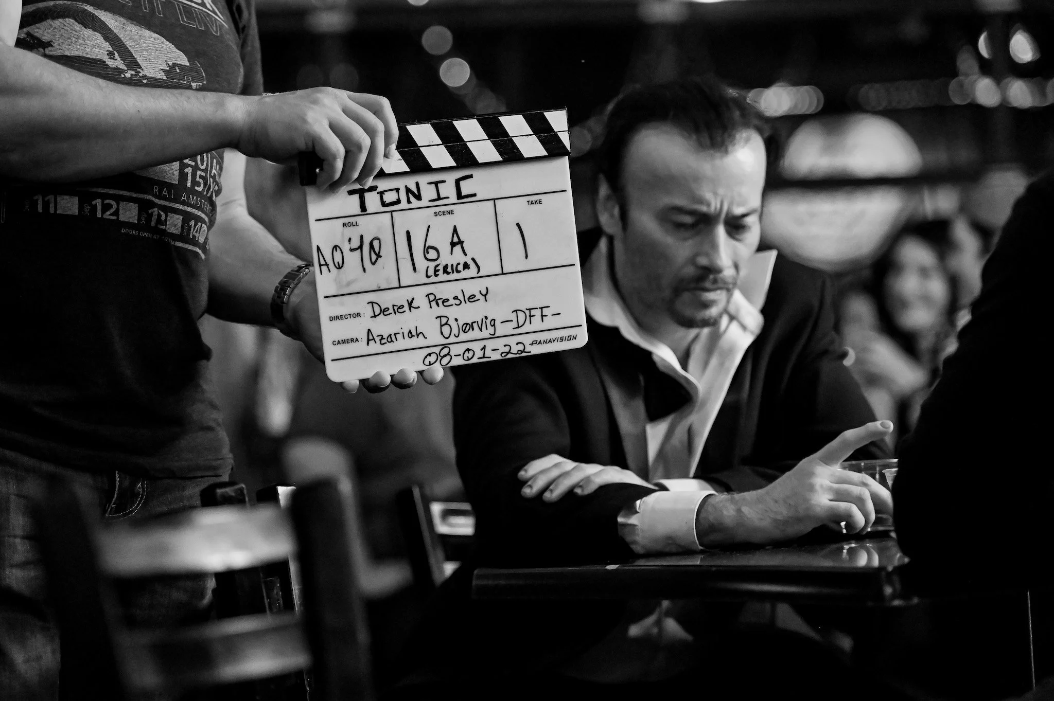 A man is holding a film slate with the word 'TONIC' written on it, on a film set. The slate displays the roll number 'A074', scene number '164', scene description '(CERICA)', and date '08-01-22'. Another man is seated at a table with his arms crossed