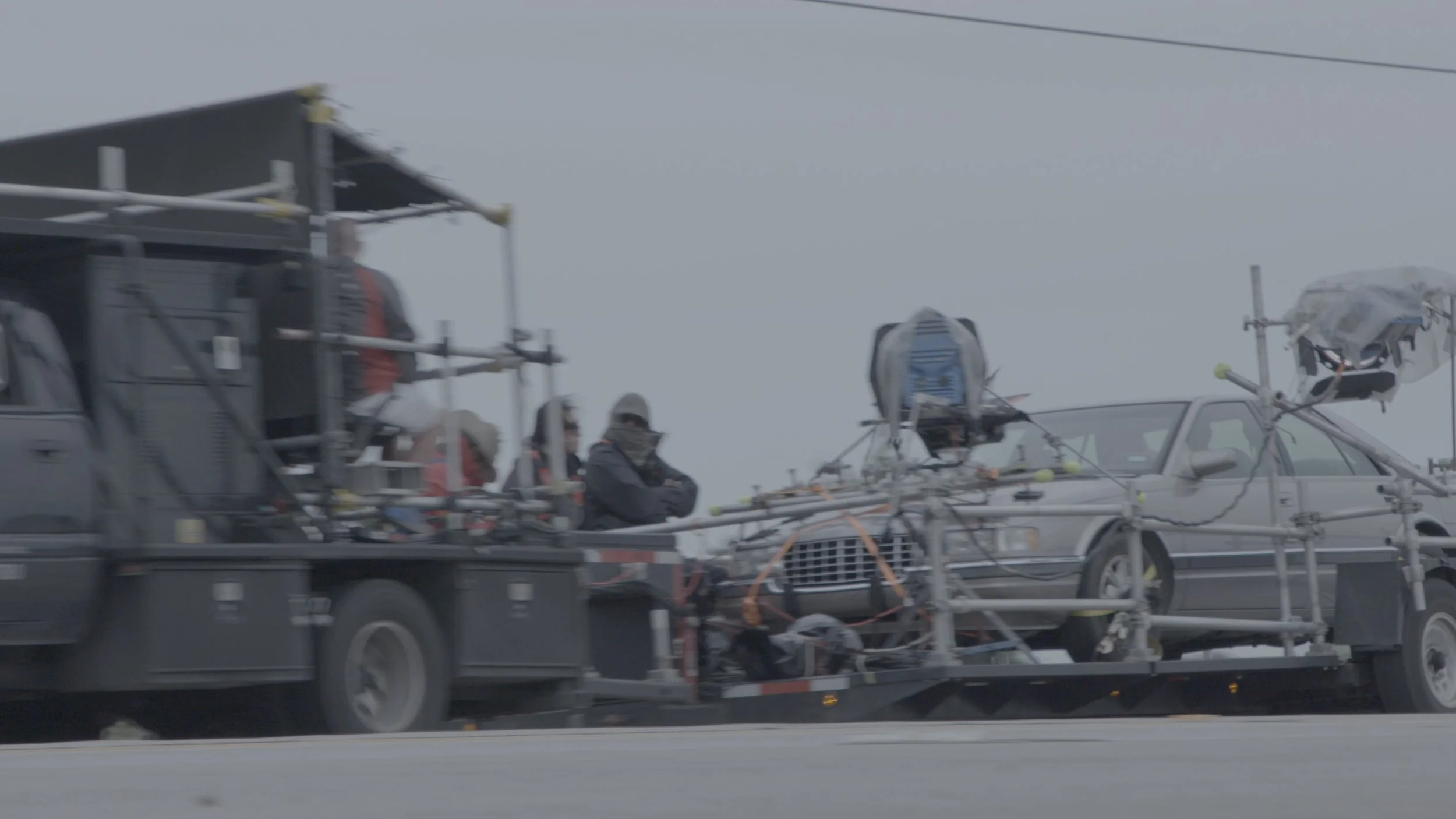 A film crew on the side of a road with equipment, cameras, and drones mounted on vehicles preparing for a shoot in cloudy weather.