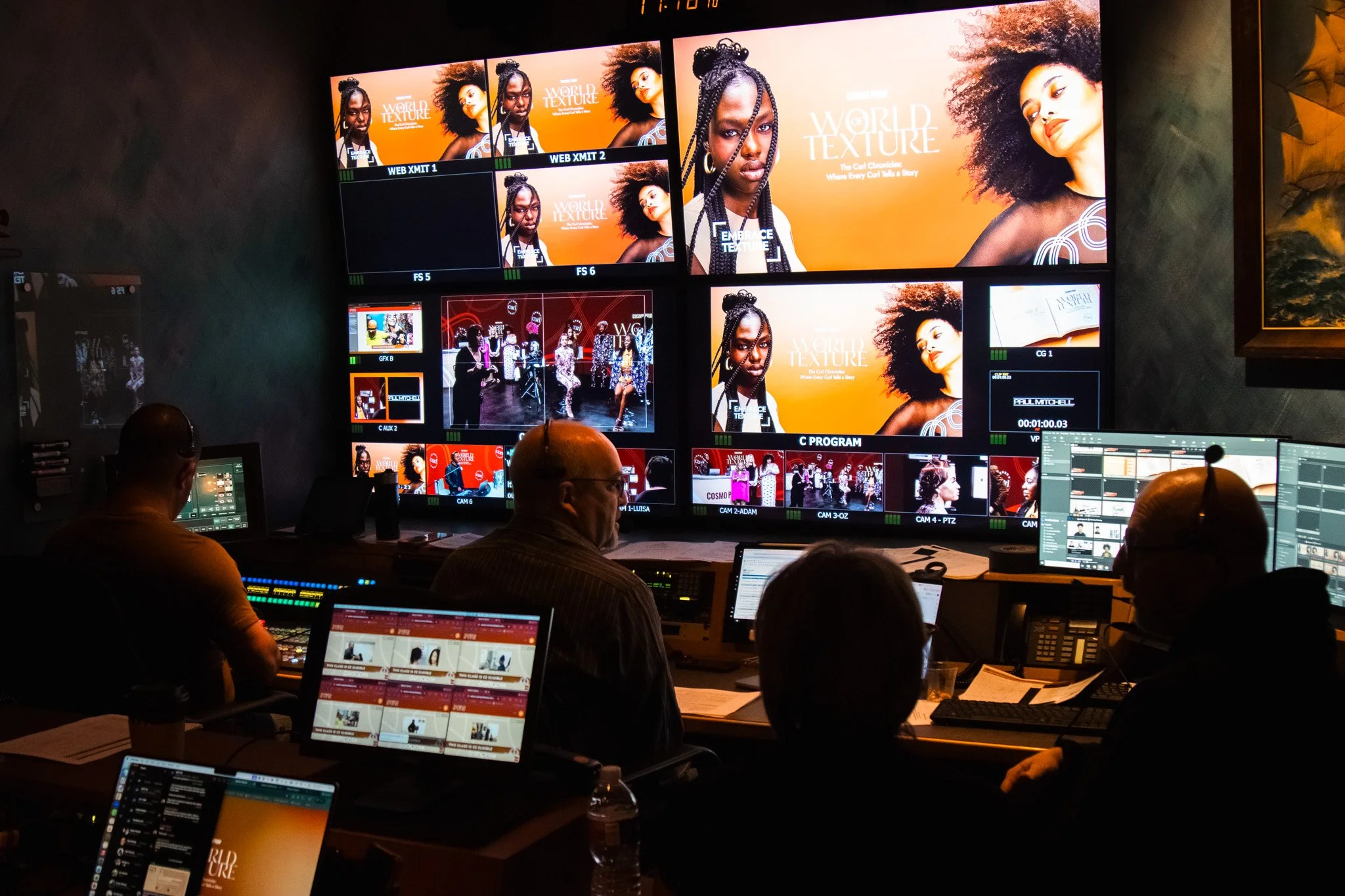 A control room with multiple screens displaying images of women with textured hair and a group of people working at their computer stations.