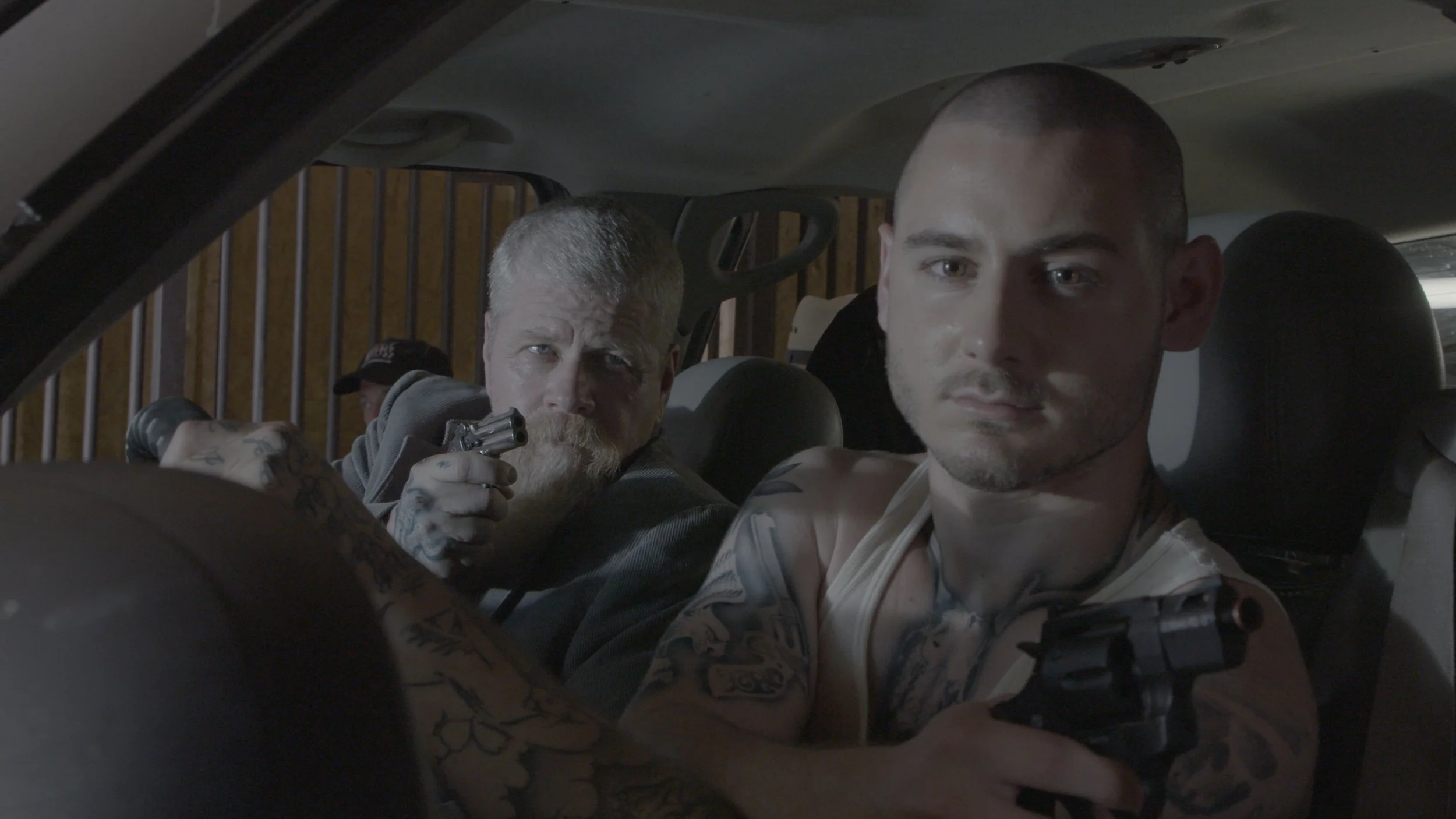 Two men are inside a vehicle. One man with tattoos and a sleeveless shirt is holding a gun and looking serious, while the other man with short hair is sitting in front with a neutral expression. The background shows a barred window and a wood wall.