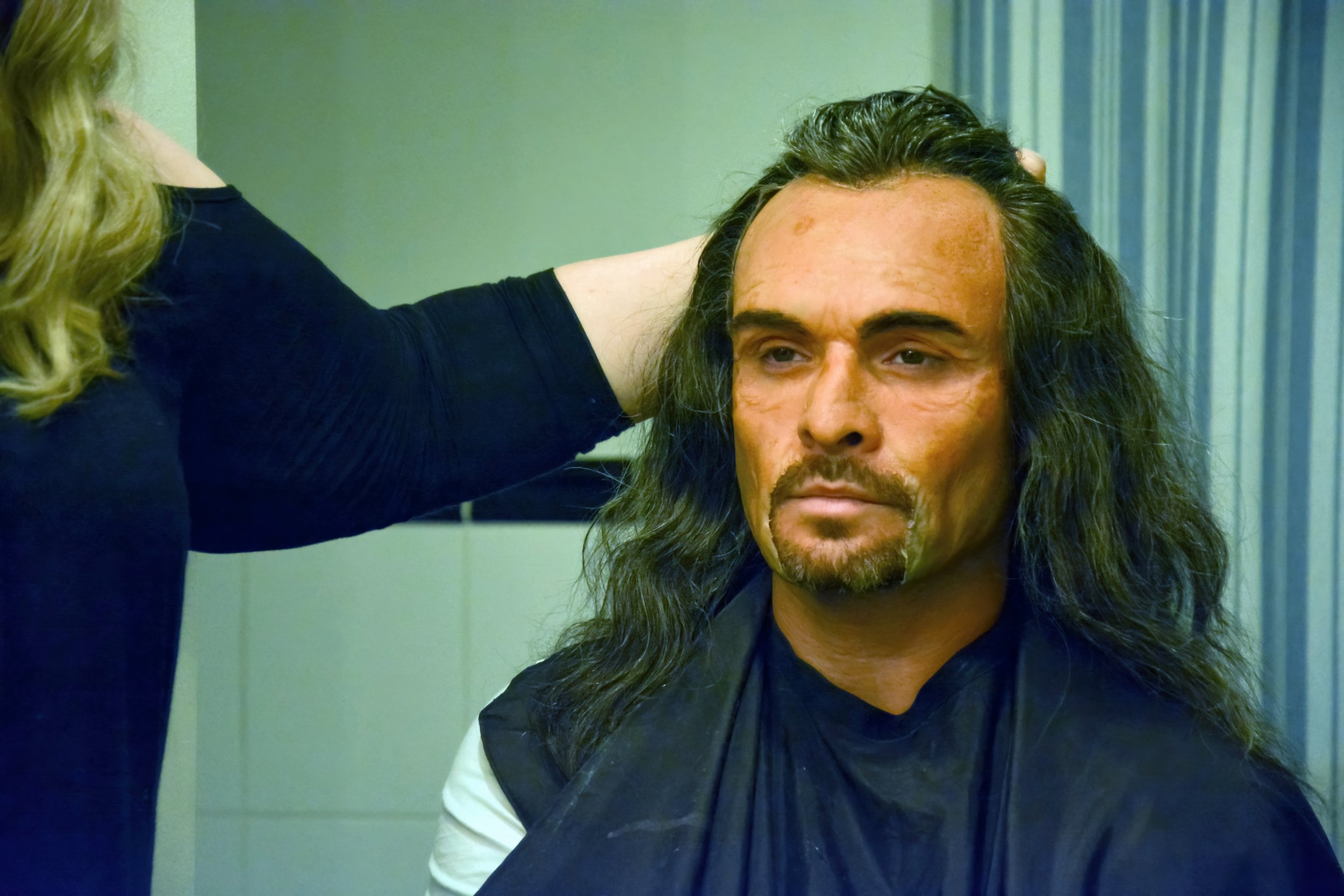 A man with long dark hair and a beard sitting in a room with a woman with curly blonde hair, who is touching his head. The man is wearing a black shirt and has a serious expression. The woman, whose face is not visible, is wearing a dark top.