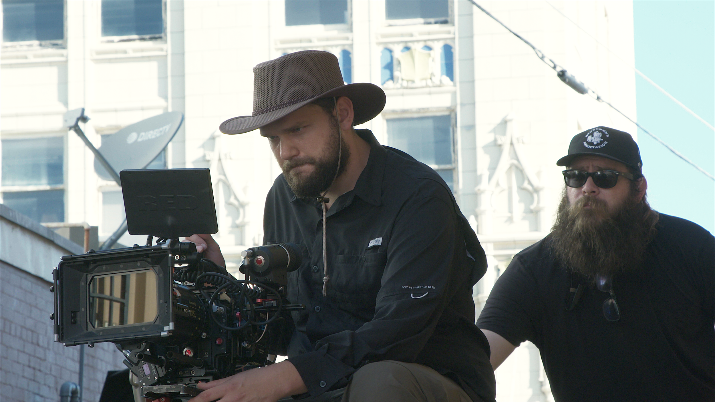 Two men on a film set outdoors, one operating a camera and the other wearing sunglasses and a hat, with city buildings and satellite dishes in the background.