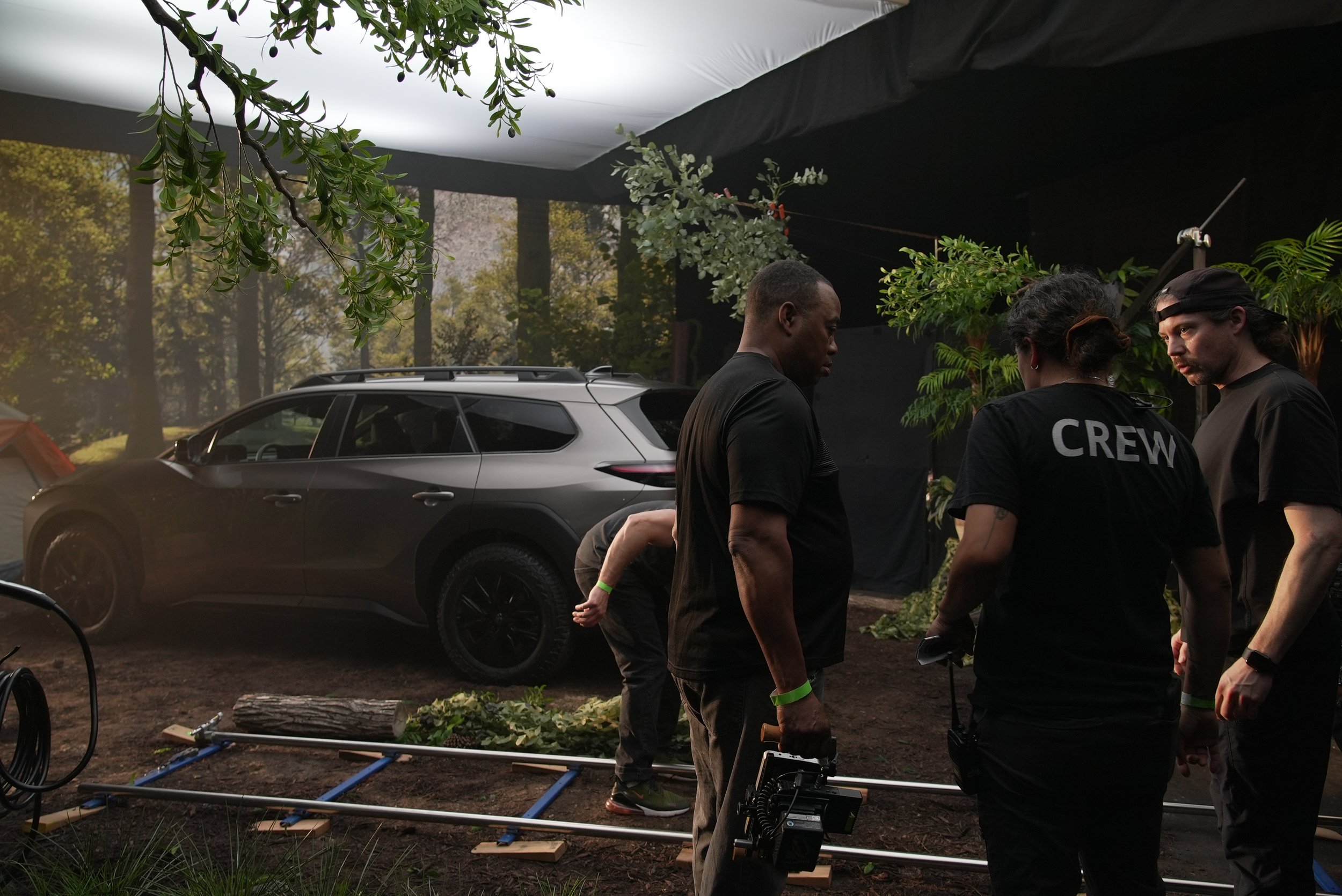 A film crew is working outdoors in a forested area with trees and clouds overhead, and a black backdrop is set up behind them. There are multiple people, some wearing black shirts with 'CREW' printed on the back, gathered around equipment including a