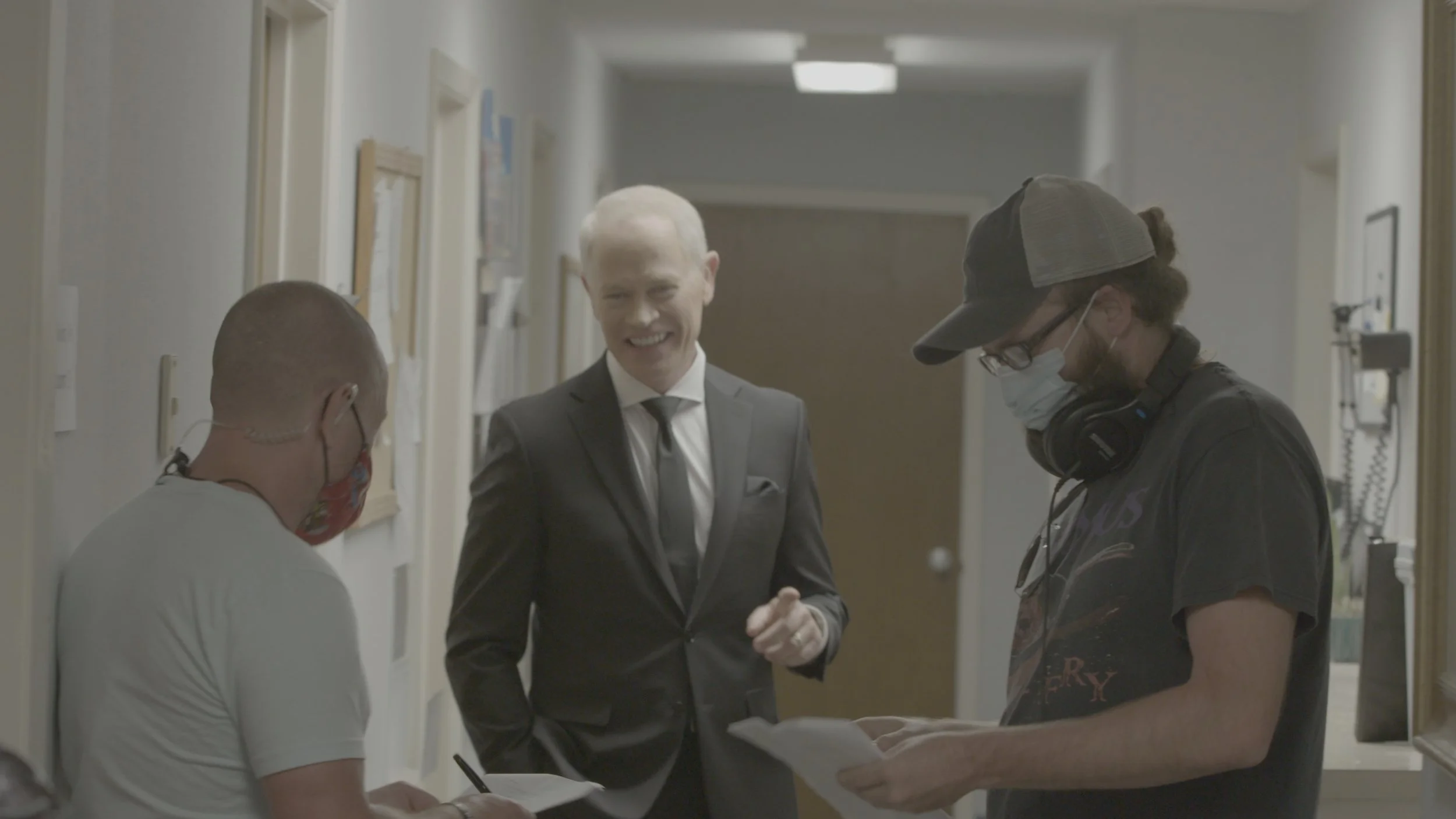 Man in a suit shaking hands with a man wearing headphones and a cap in a hospital hallway, with two other masked individuals nearby.