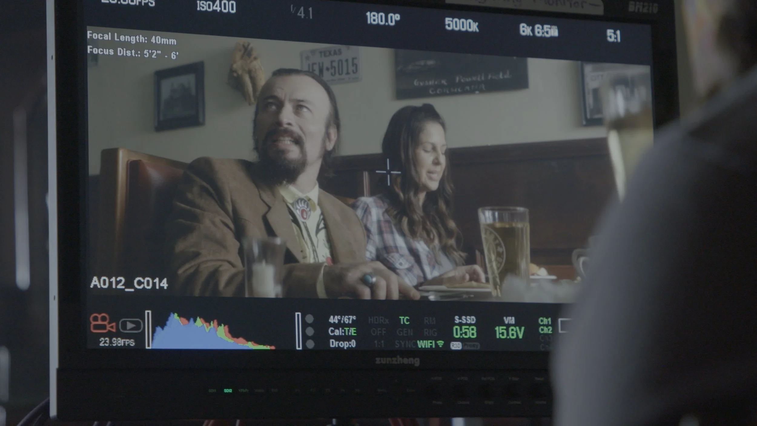 A scene from a film or television show seen through a camera monitor, featuring a man with long hair and a beard and a woman with long dark hair, sitting at a restaurant table with drinks, with wall decorations including framed pictures and a mounted