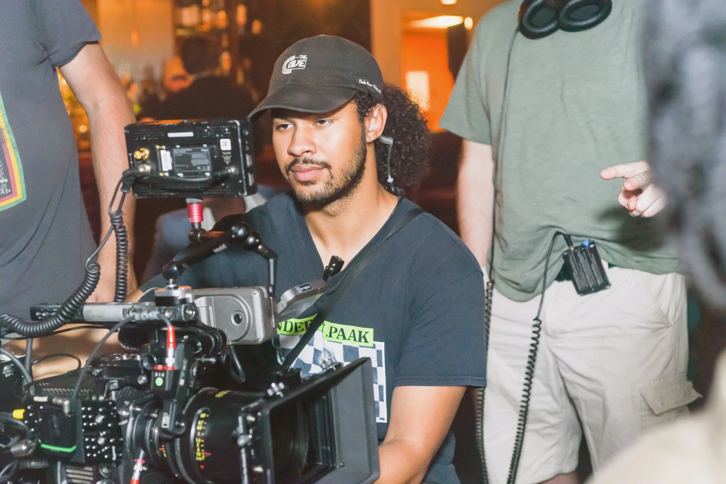 A man with curly hair and a beard, wearing a black cap and a black t-shirt, is sitting in front of a professional camera setup, surrounded by crew members on a film set.