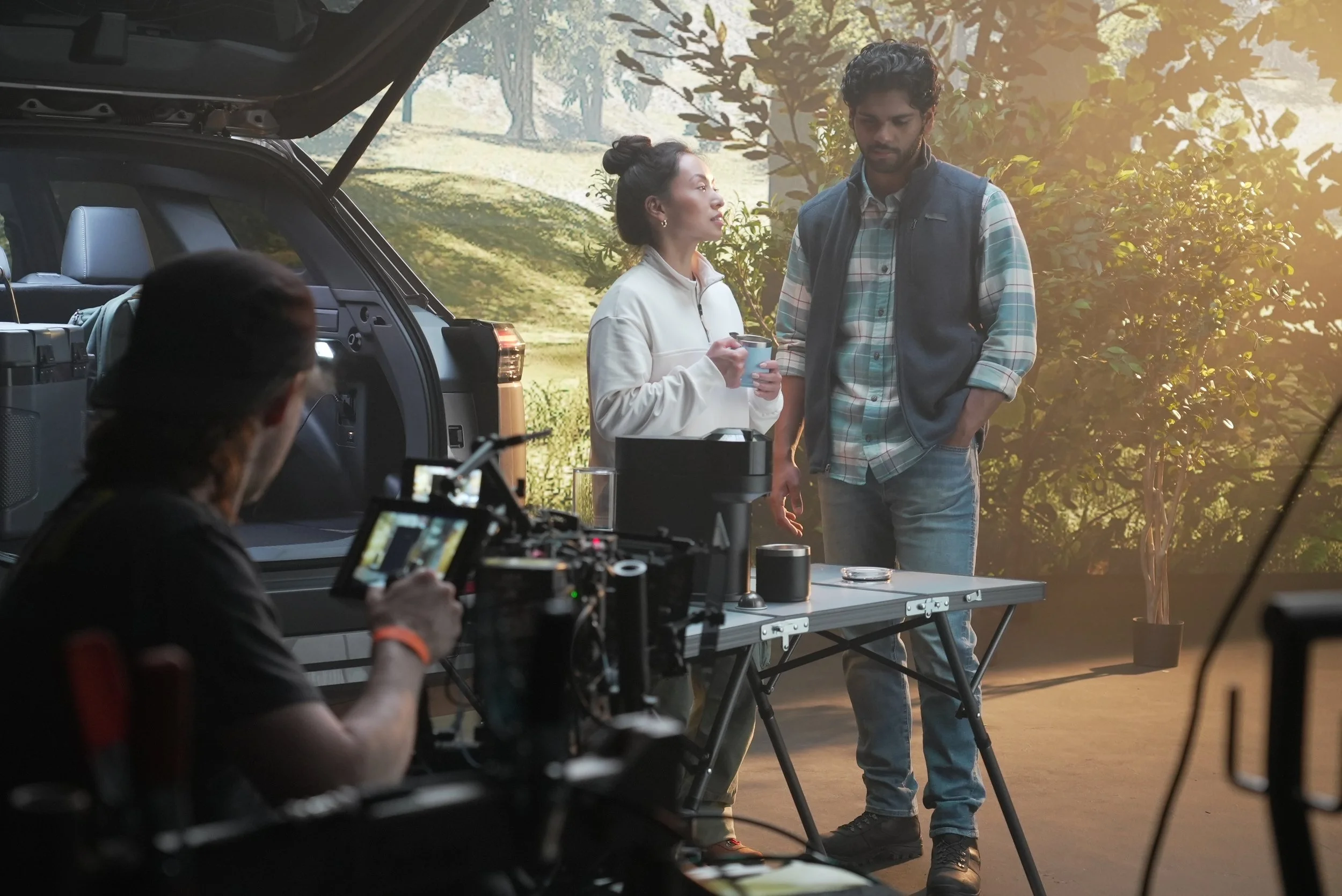 Behind the scenes of the Toyota Bz Woodland shoot at AMS Pictures, with a crew member operating a camera, two actors in discussion by a vehicle with an open trunk outdoors, surrounded by trees and foliage, during daytime.