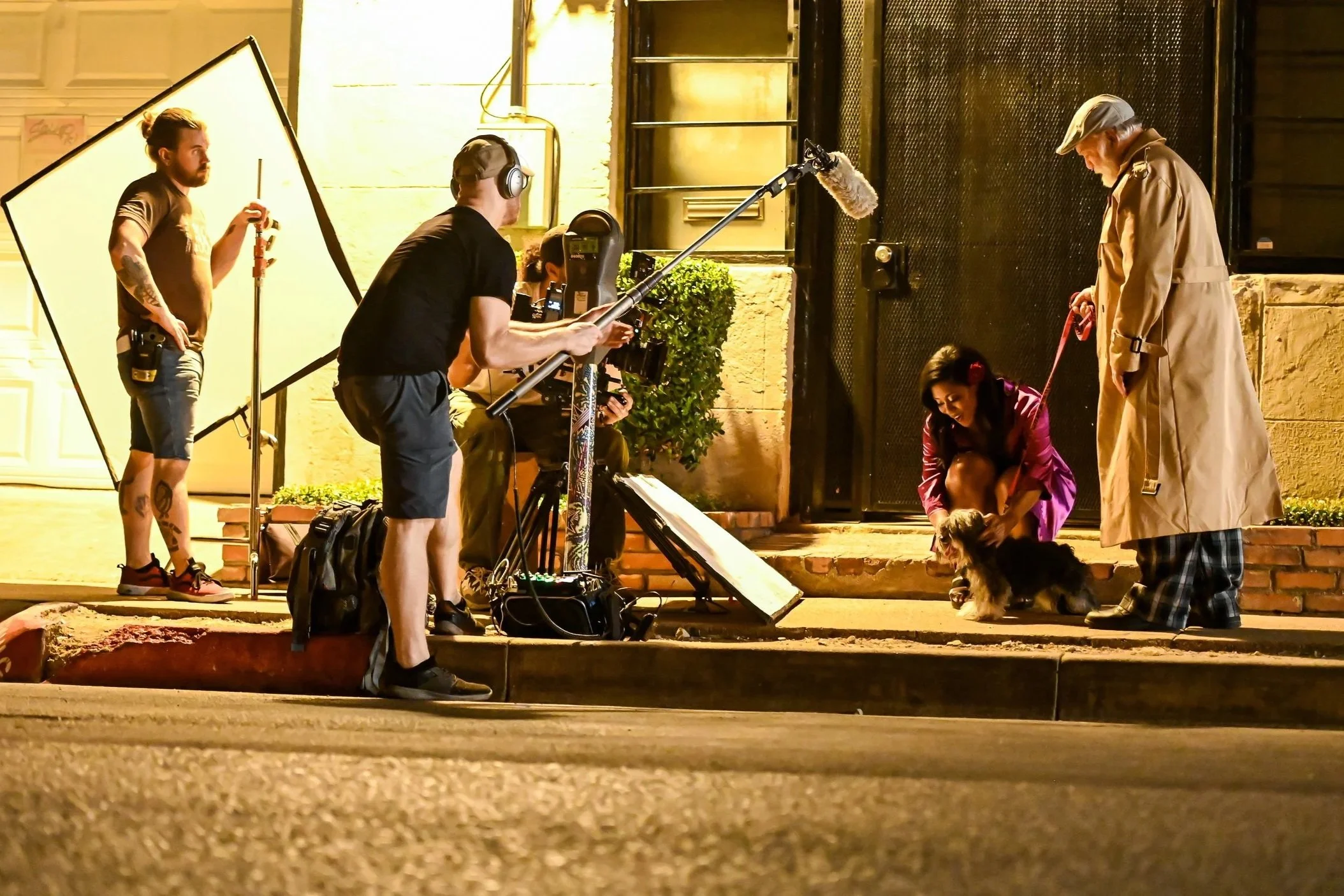 Tonic film production crew setting up shots on a front porch at night, with actress Aimee Masterson kneeling next to a small dog and actor Richard Riehle holding the dog's leash.