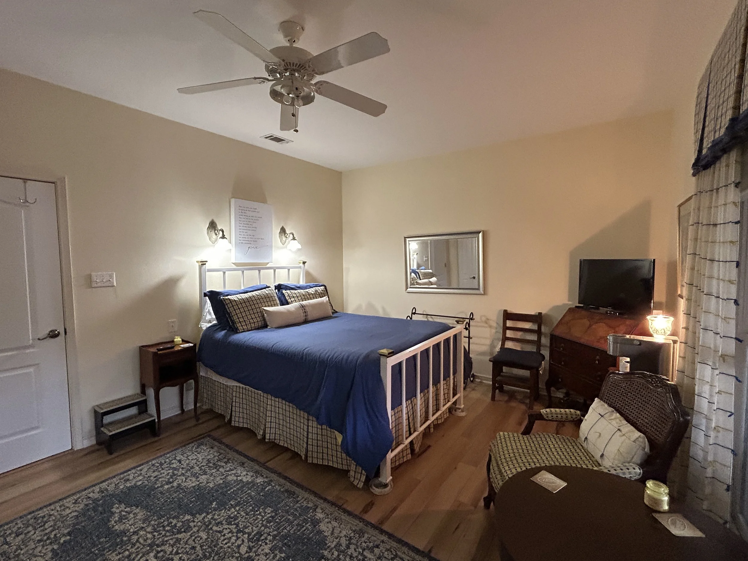 A cozy bedroom with a bed dressed in blue bedding and plaid pillows, a ceiling fan, a mirror on the wall, a small wooden chair, a dresser with a TV and a lamp, a cushioned armchair with pillows, and a window with patterned curtains.