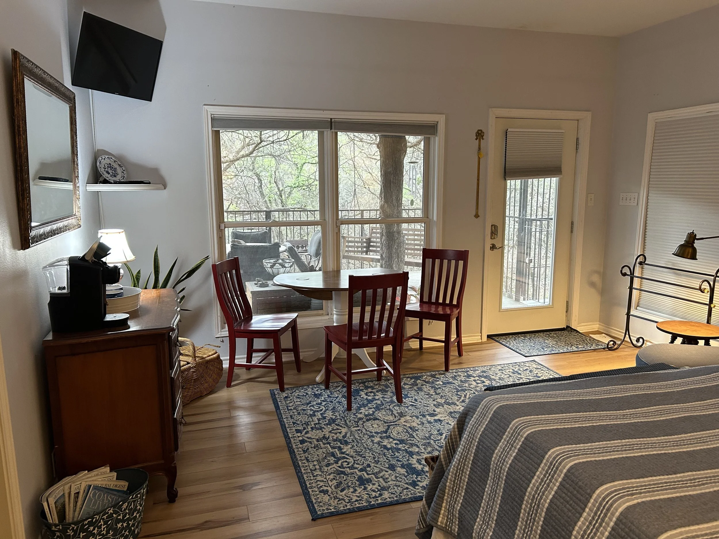Living room with window view of a balcony, a circular dining table with four red chairs, a bed with striped bedding, a small wooden side table, a door with a window, and a flat-screen TV mounted on the wall.