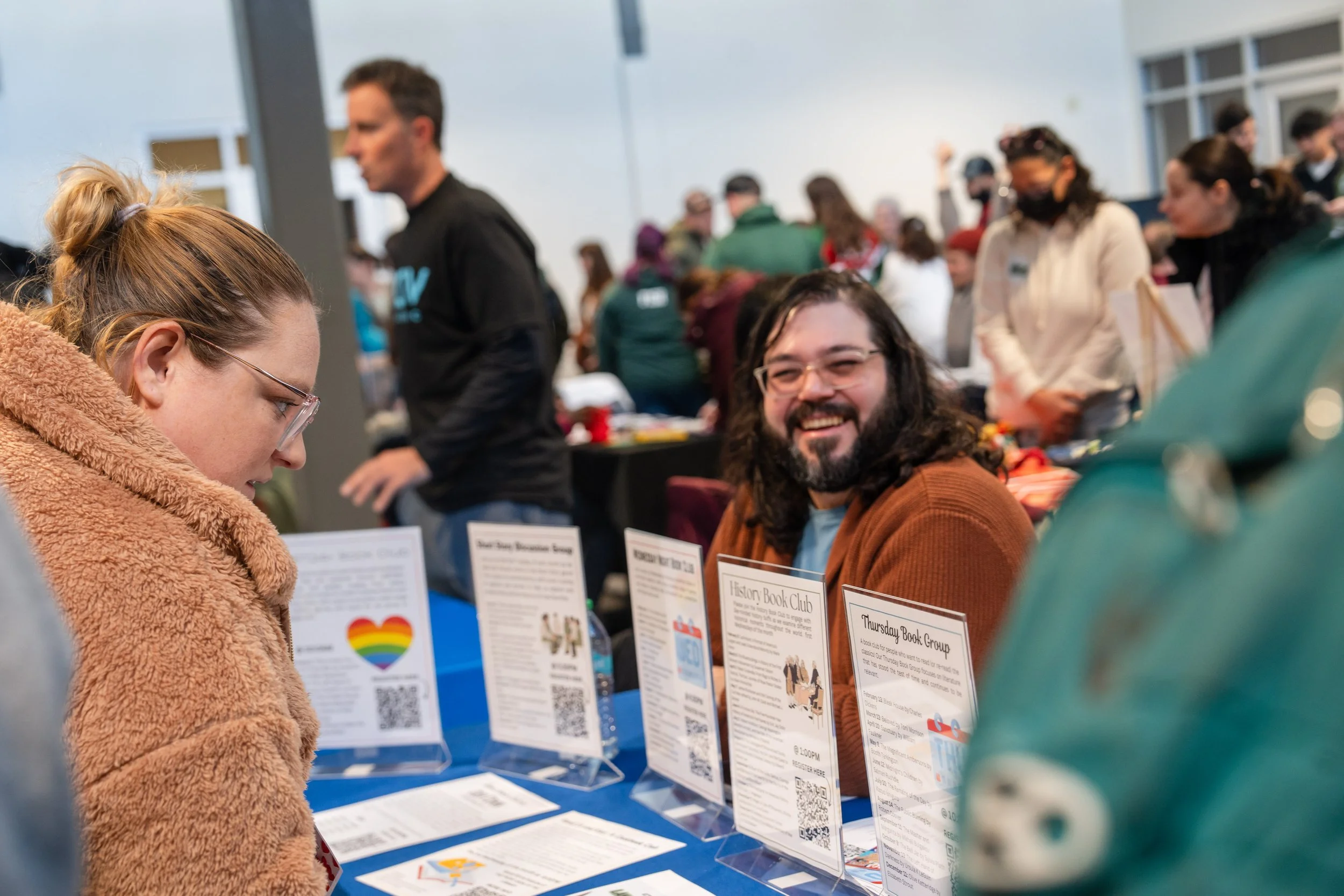 A woman with glasses and a tan fuzzy coat looking at informational flyers on a table. A smiling man with glasses, a beard, and long dark hair sits across from her, with tables and many people in the background.