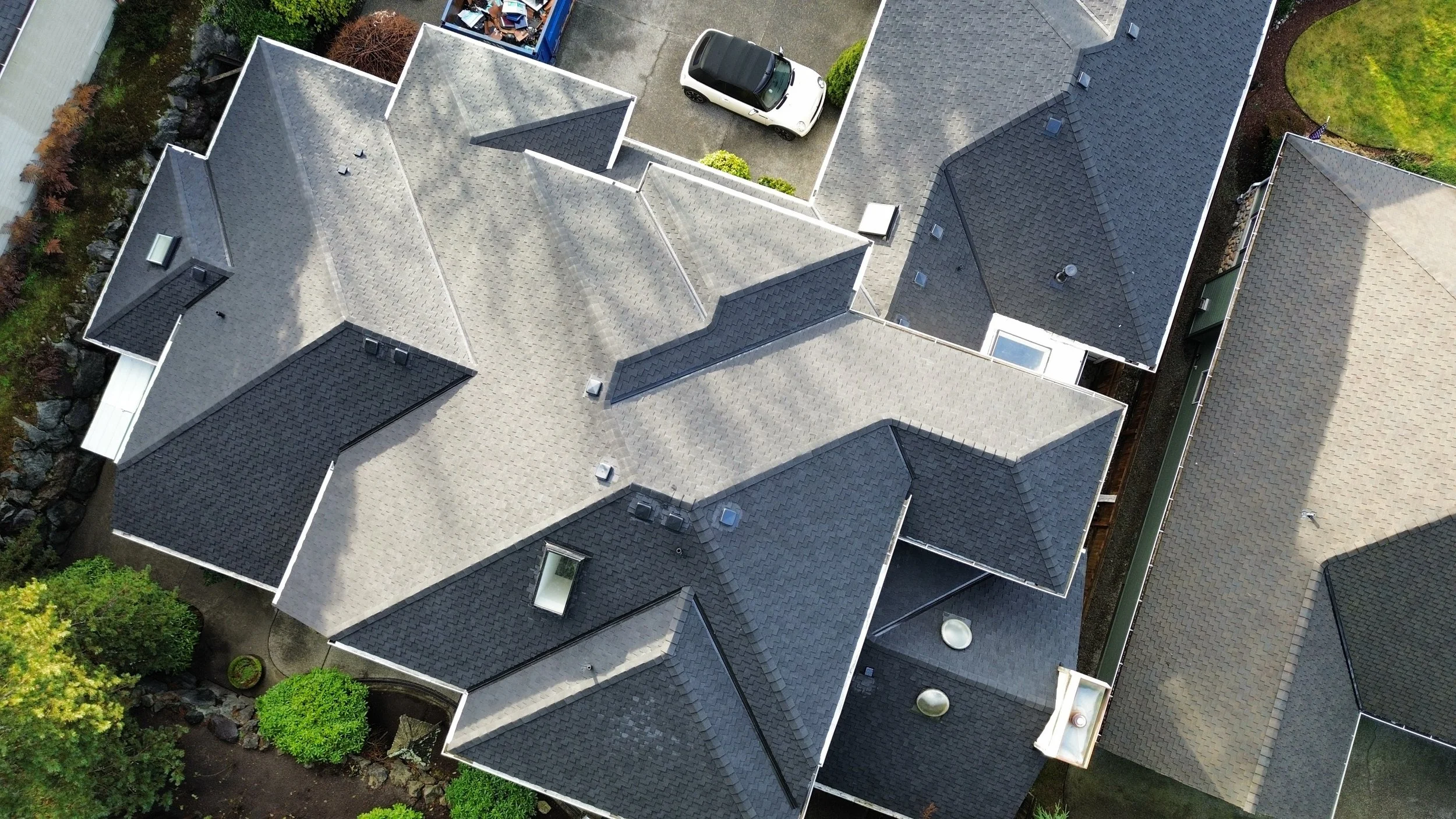 An aerial view of a residential neighborhood showing the rooftops of several houses with various roof styles and colors. There is a small parking area with a white car and some greenery around.