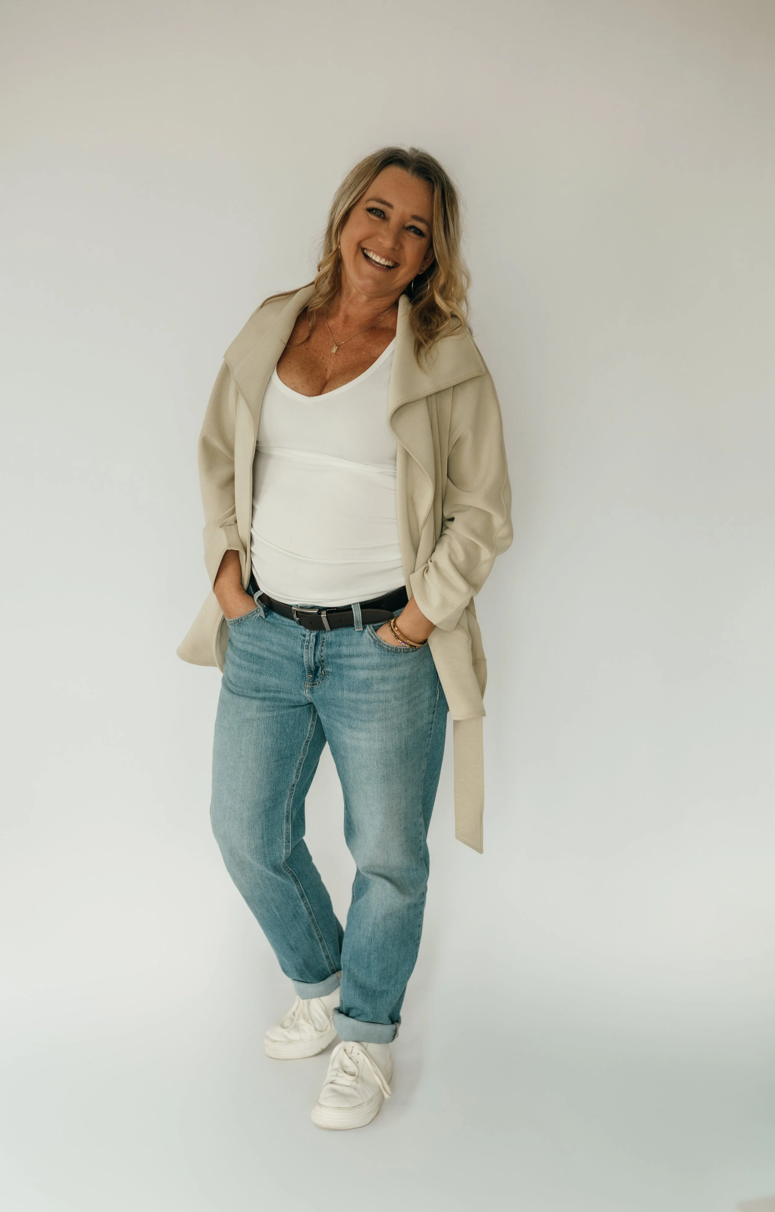 A woman with blonde hair wearing a beige coat, white top, light blue jeans, and white sneakers, smiling and standing against a plain white background.