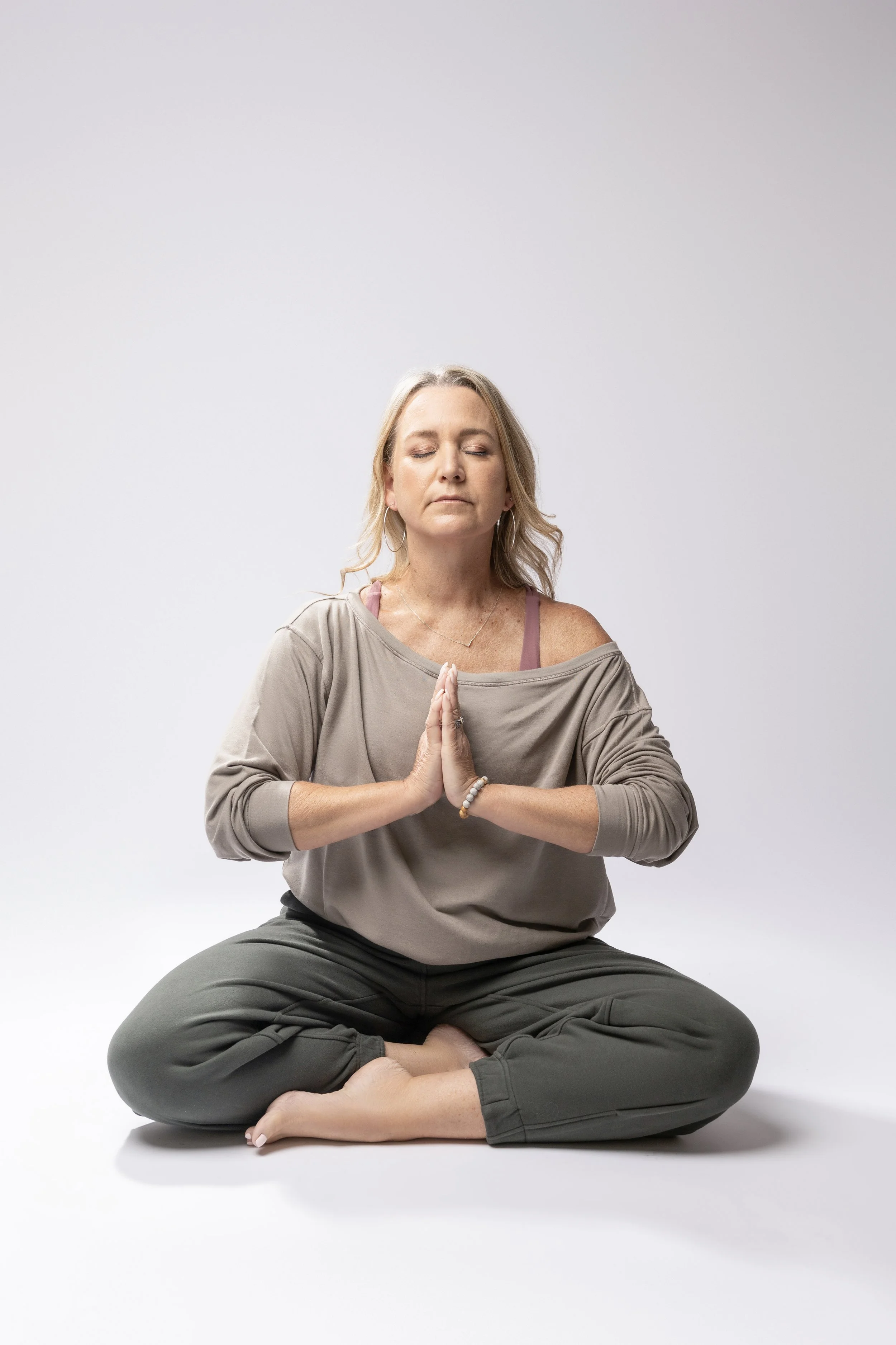 A woman practicing yoga in a seated meditation pose with her hands in prayer position and closed eyes in a minimalist studio.