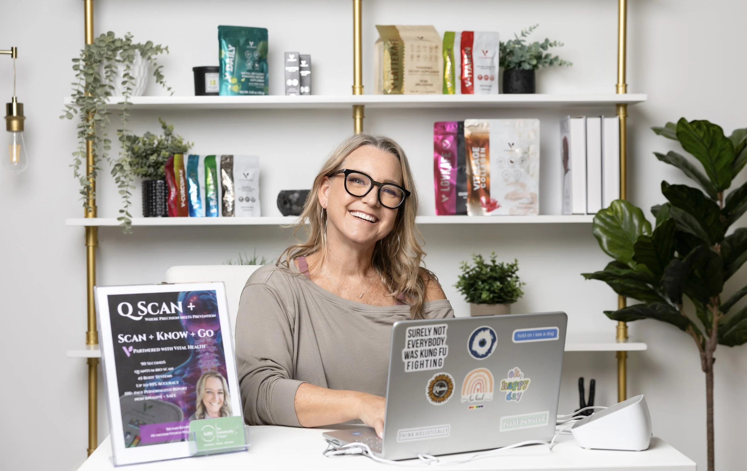 A smiling woman sitting at a desk with a laptop covered in stickers, in a modern office with white shelves, plants, and promotional posters.