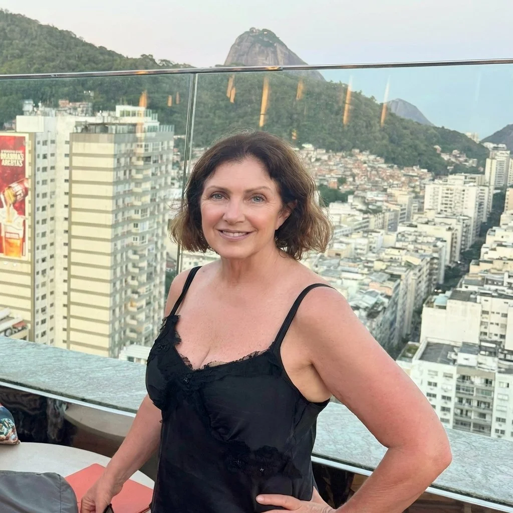 A smiling woman in a black spaghetti strap top standing on a rooftop or balcony with a view of a cityscape and mountains in the background.