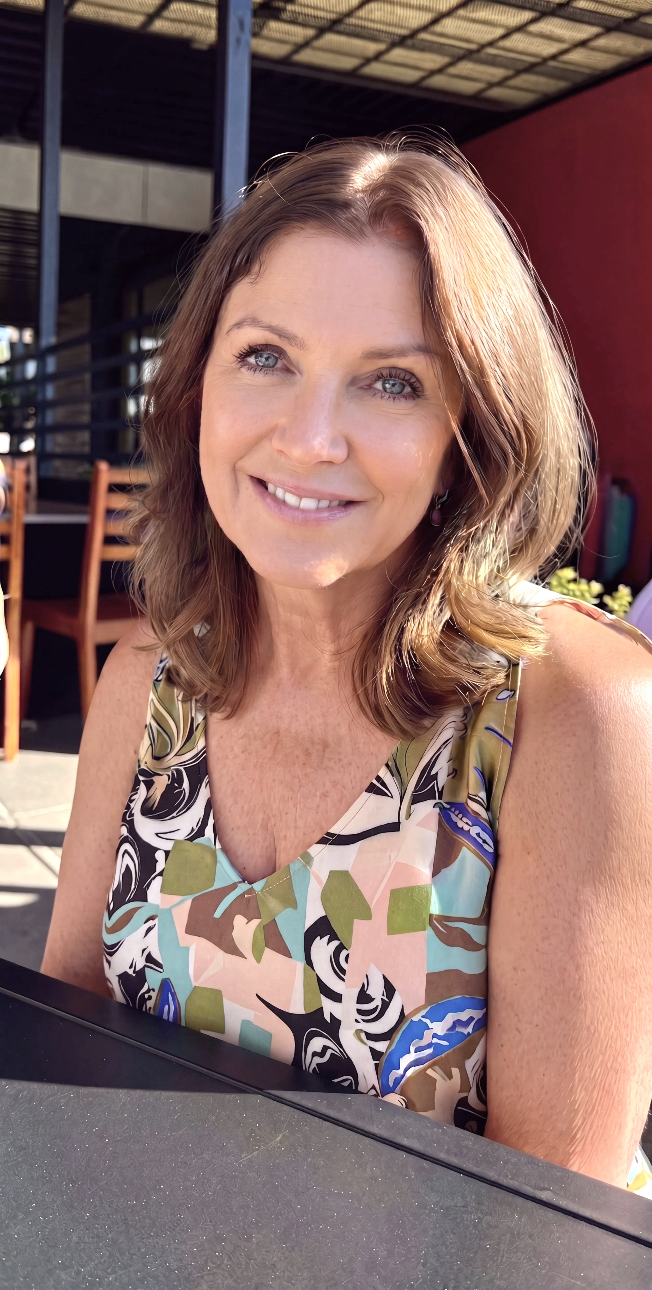 A woman with shoulder-length wavy light brown hair, blue eyes, and light skin, smiling in sunlight, wearing a sleeveless dress with a colorful, abstract pattern, sitting outdoors at a table.