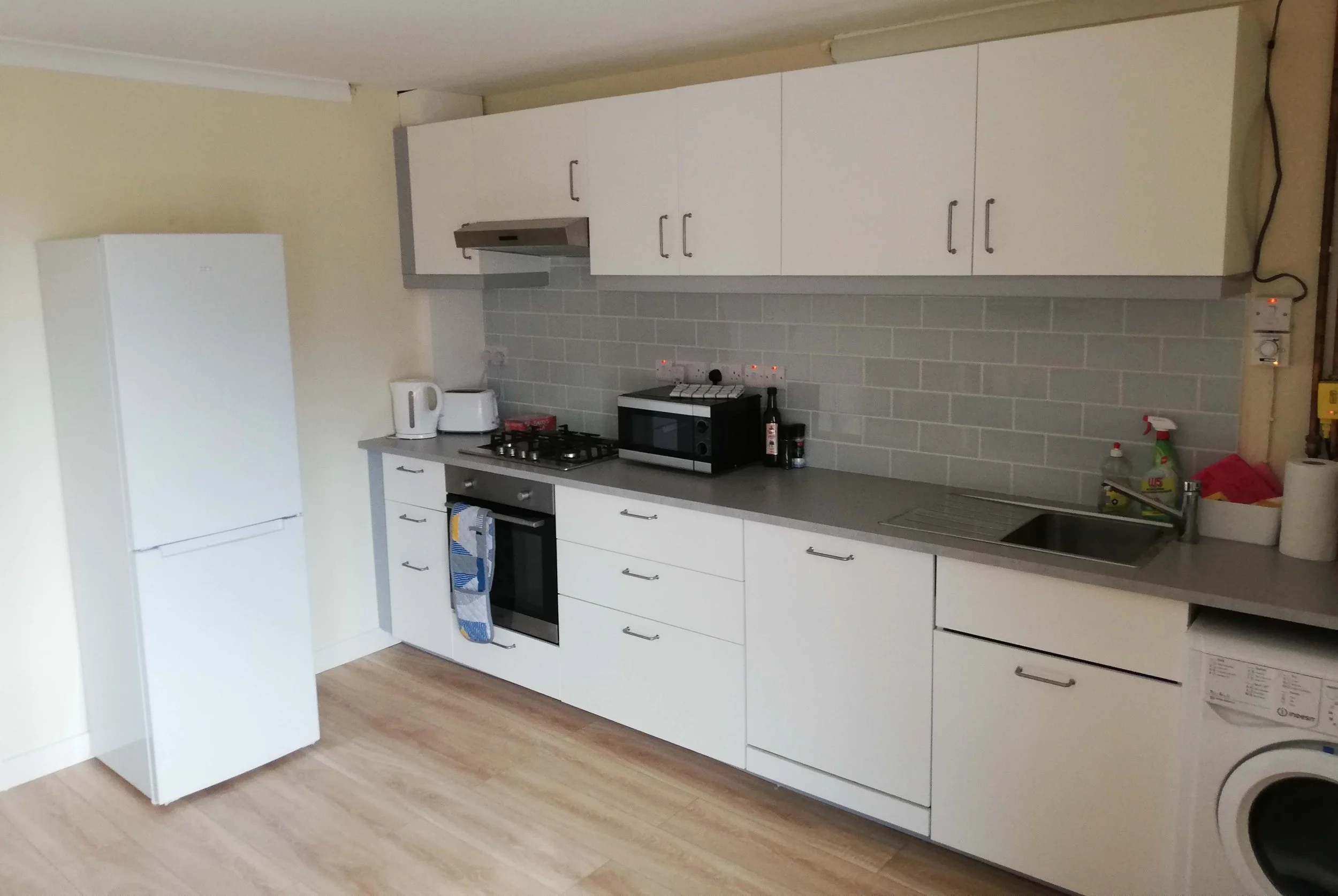 Kitchen with white cabinets, gray countertop, refrigerator, oven, microwave, kettle, toaster, stove, and sink against a wall with gray tile backsplash.