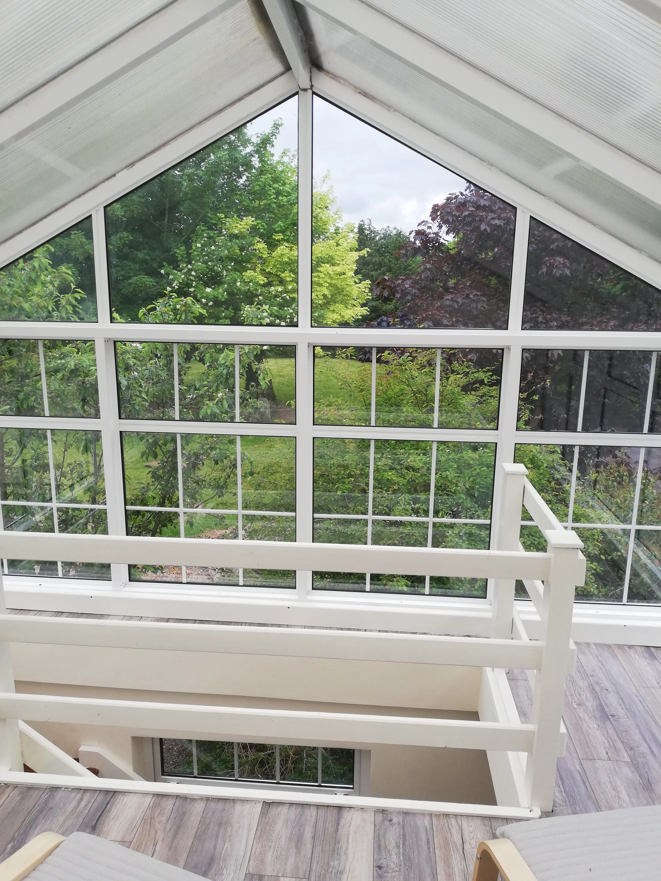 View from inside a conservatory showing large glass windows with a garden and trees outside, with wooden floors and part of a staircase railing.