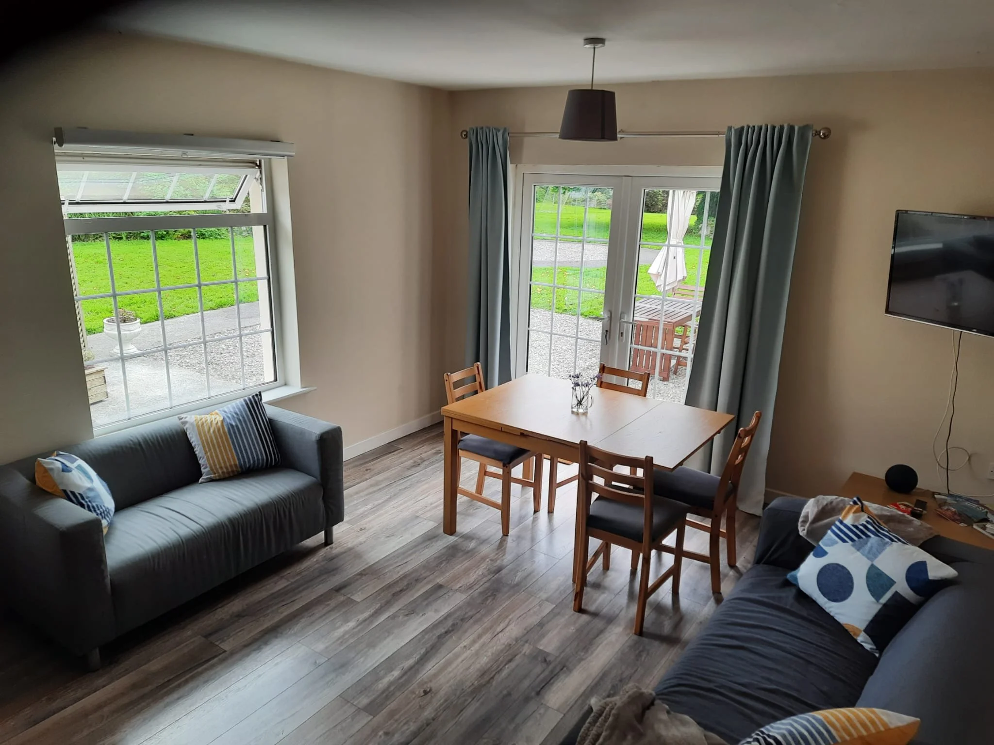 A living room with two sofas, a wooden dining table with four chairs, a flat-screen TV on the wall, a window, and a glass sliding door leading to an outdoor patio with a table, chairs, and an umbrella. The room has wood flooring and light-colored walls.