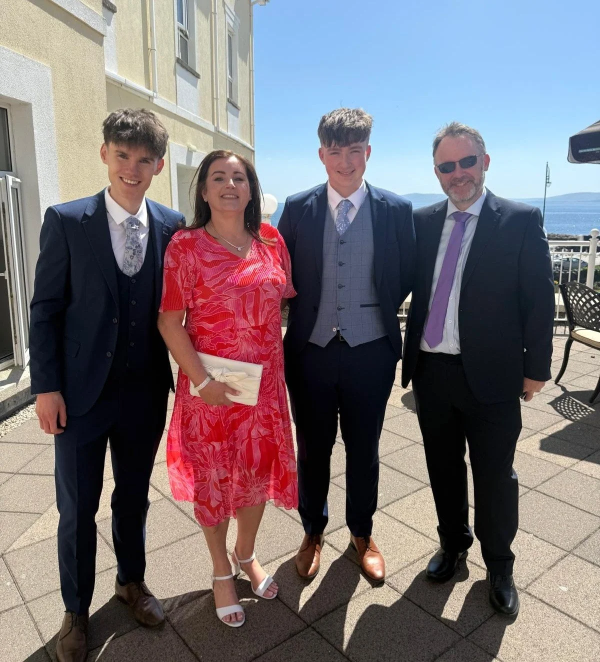 A family standing on a balcony overlooking the ocean, dressed in formal attire, smiling for a photo on a sunny day.