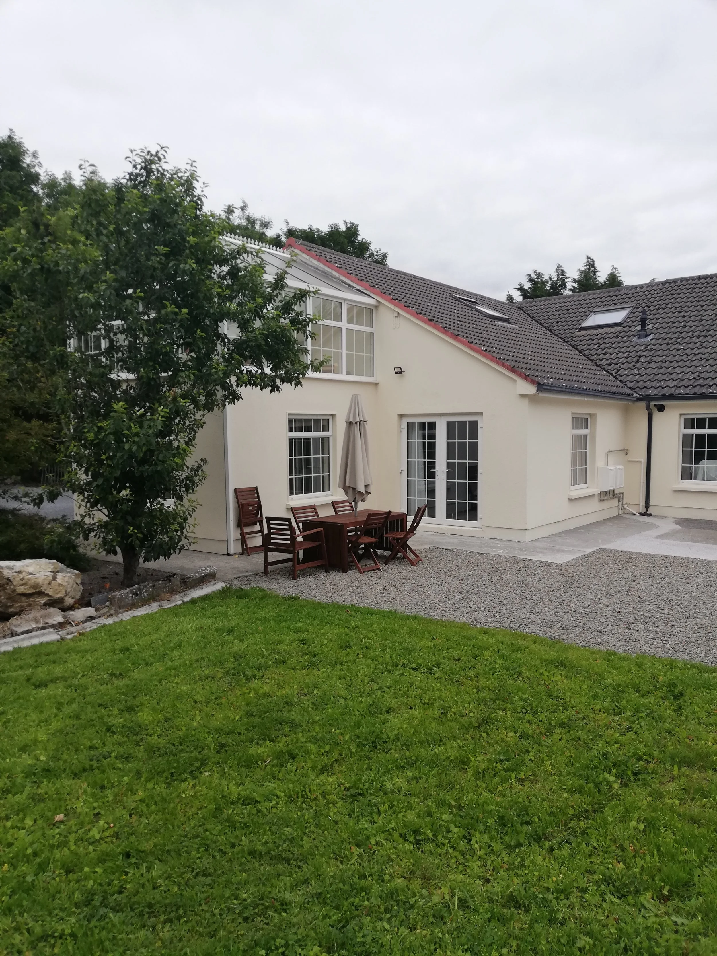 A backyard view of a white house with a patio, green grass, and a tree on the left. There is an outdoor dining table with chairs and an umbrella.