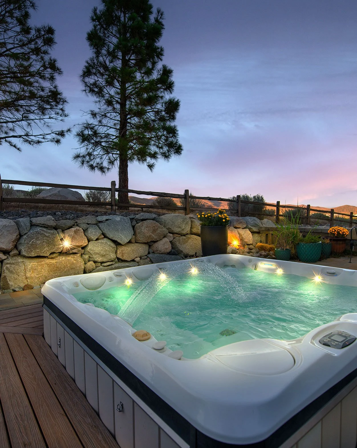 A hot tub on a wooden deck outdoors at sunset with water jets on, surrounded by potted plants, a stone wall, and a wooden fence. In the background, there is a tall pine tree and scenic desert landscape with mountains and a twilight sky.