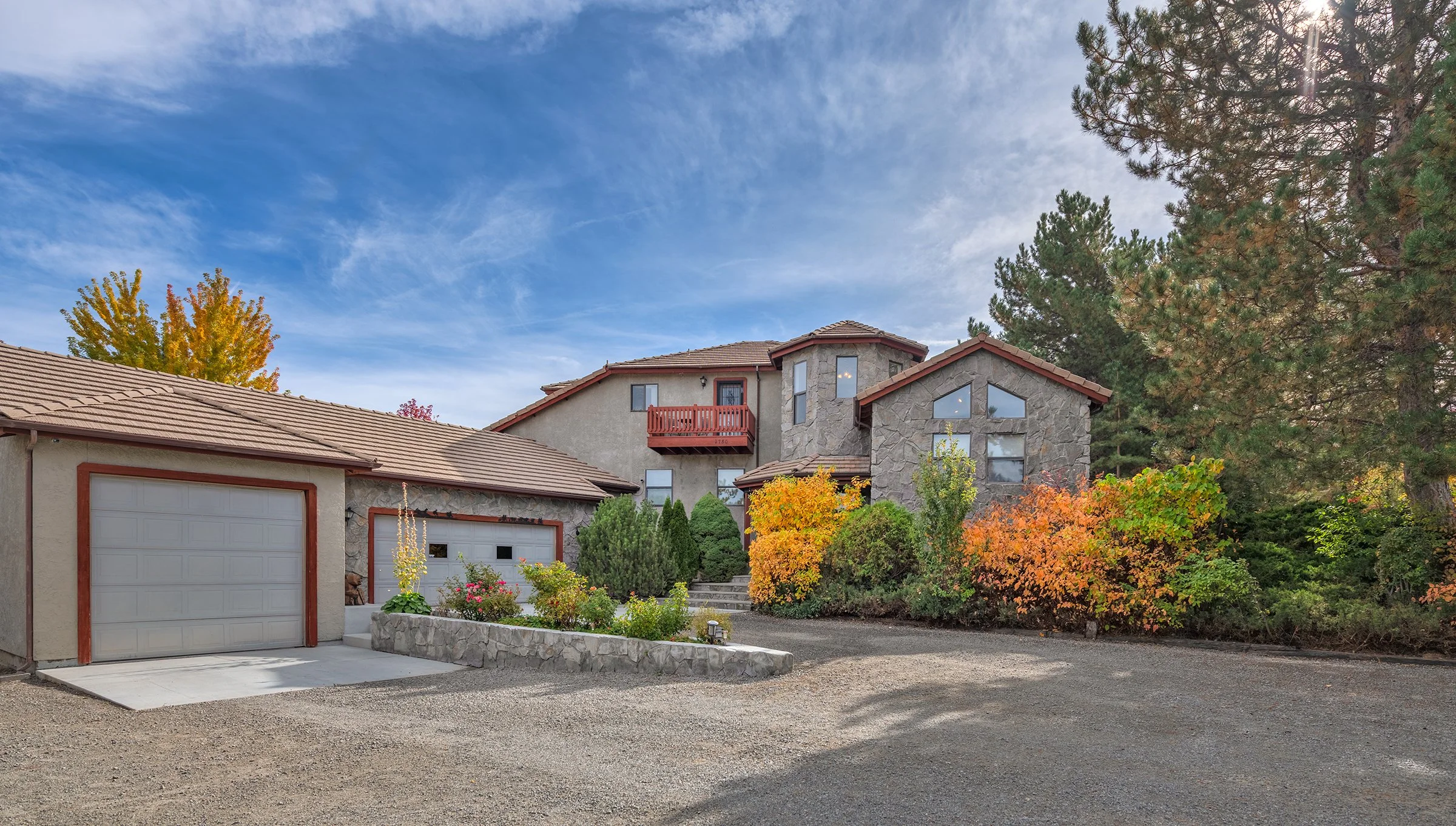 A large house with a red-tiled roof, stone and stucco exterior, and multiple windows surrounded by trees with fall foliage. The house has a driveway and a garden bed with flowering plants and bushes.