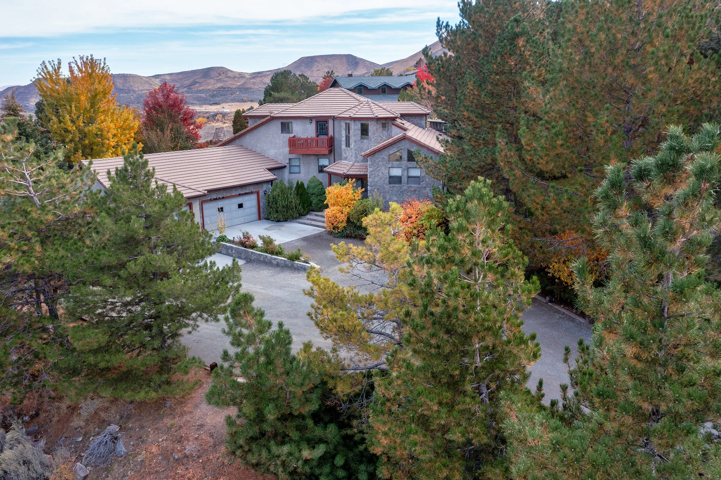 A large, multi-story house surrounded by tall pine trees, with a driveway, garage, and autumn foliage in a mountainous landscape.