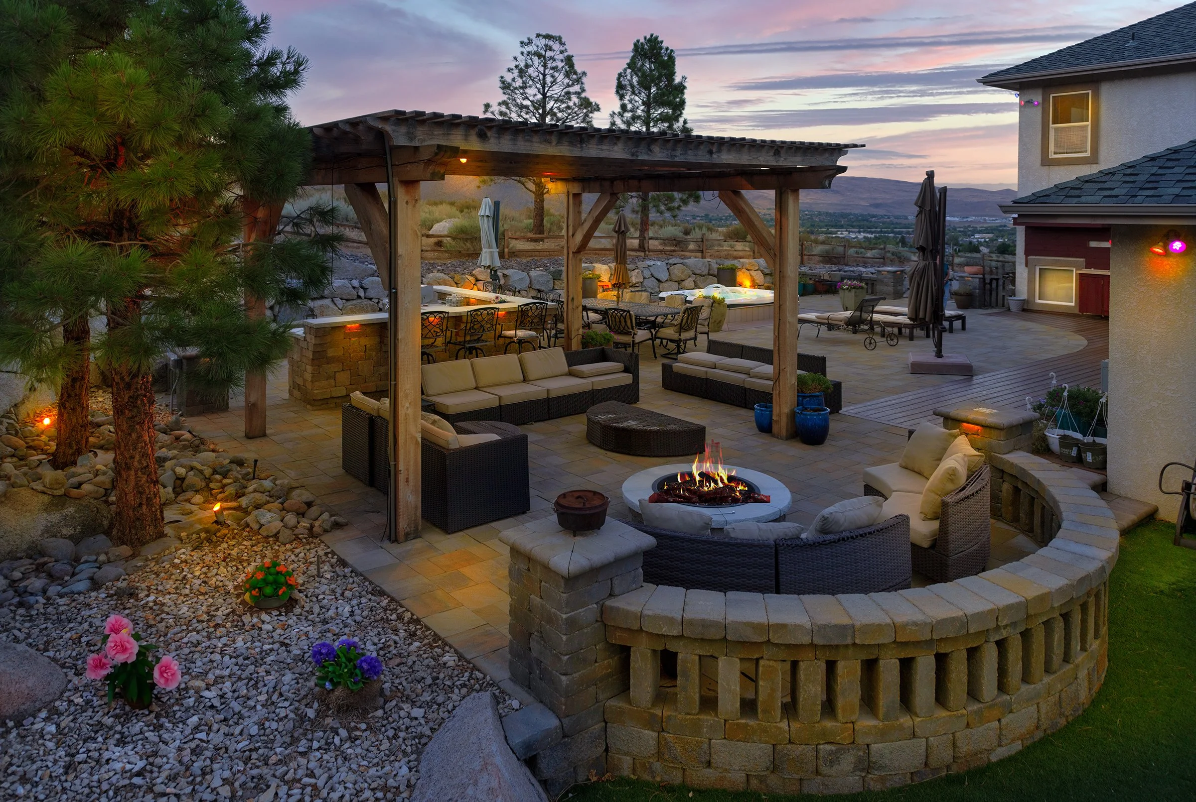 A backyard patio area during sunset with outdoor furniture, a fire pit, and a hot tub, surrounded by trees, rocks, and landscaping.