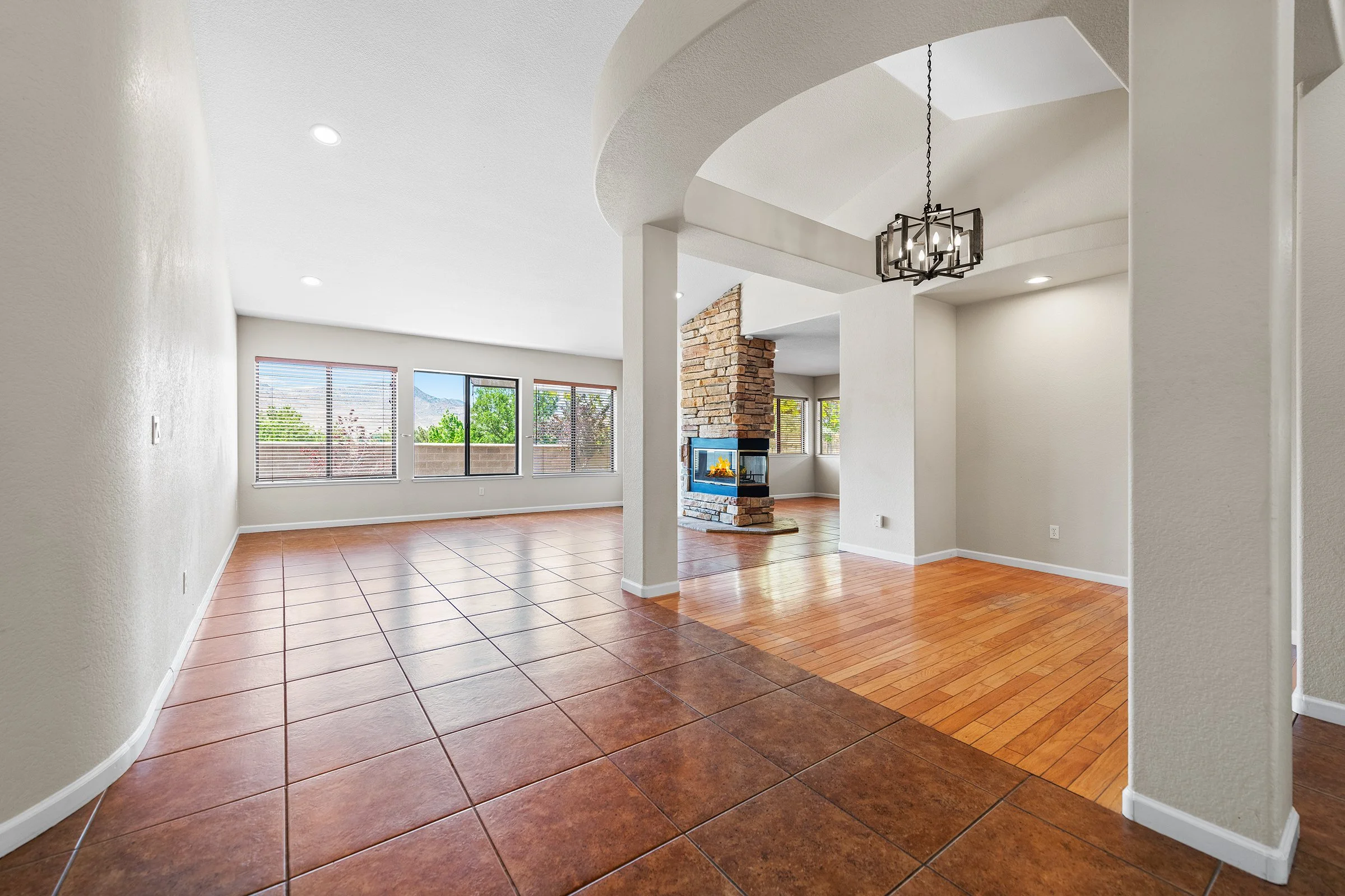 Empty living room with large windows, white walls, terracotta floor tiles, hardwood area, stone fireplace, and modern chandelier.