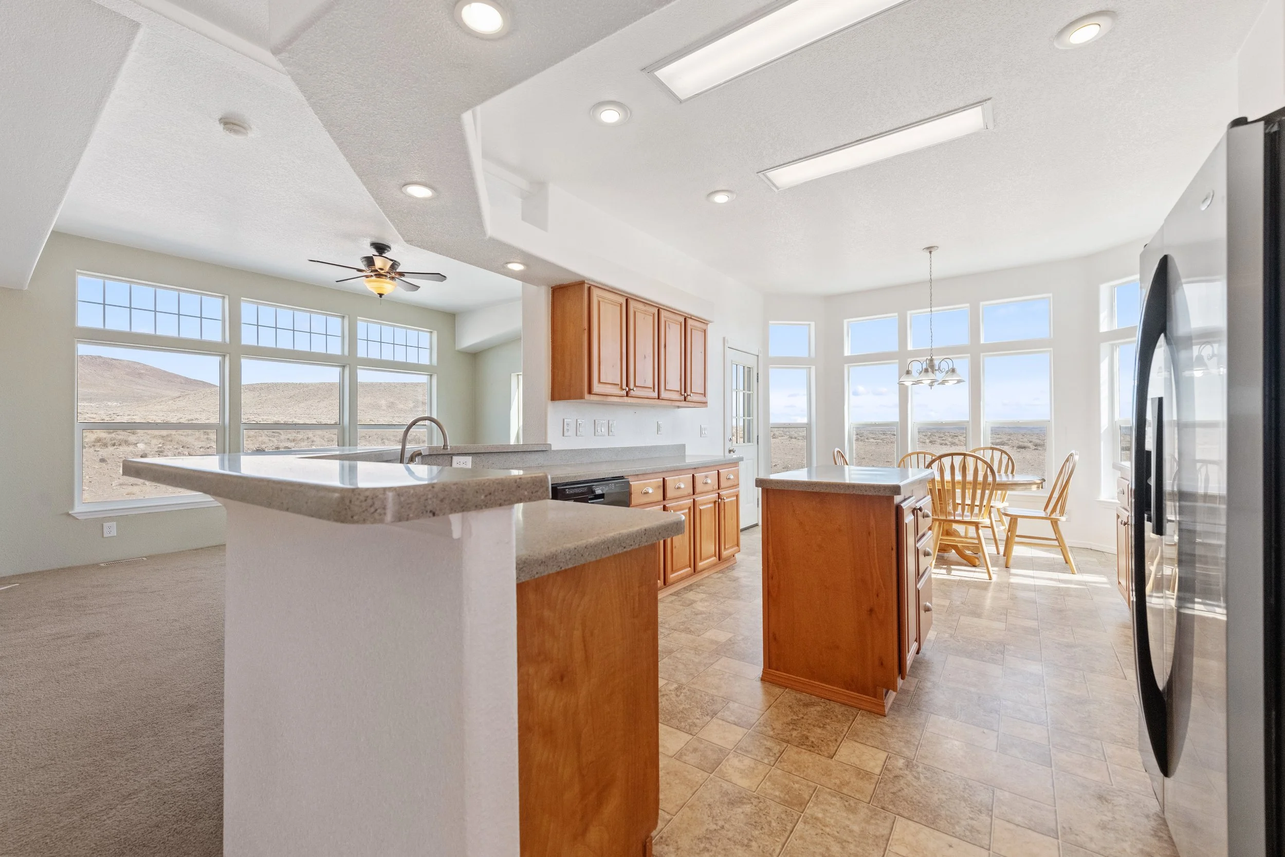 Bright open kitchen and dining area with large windows, wooden cabinets, tiled flooring, and a breakfast bar.