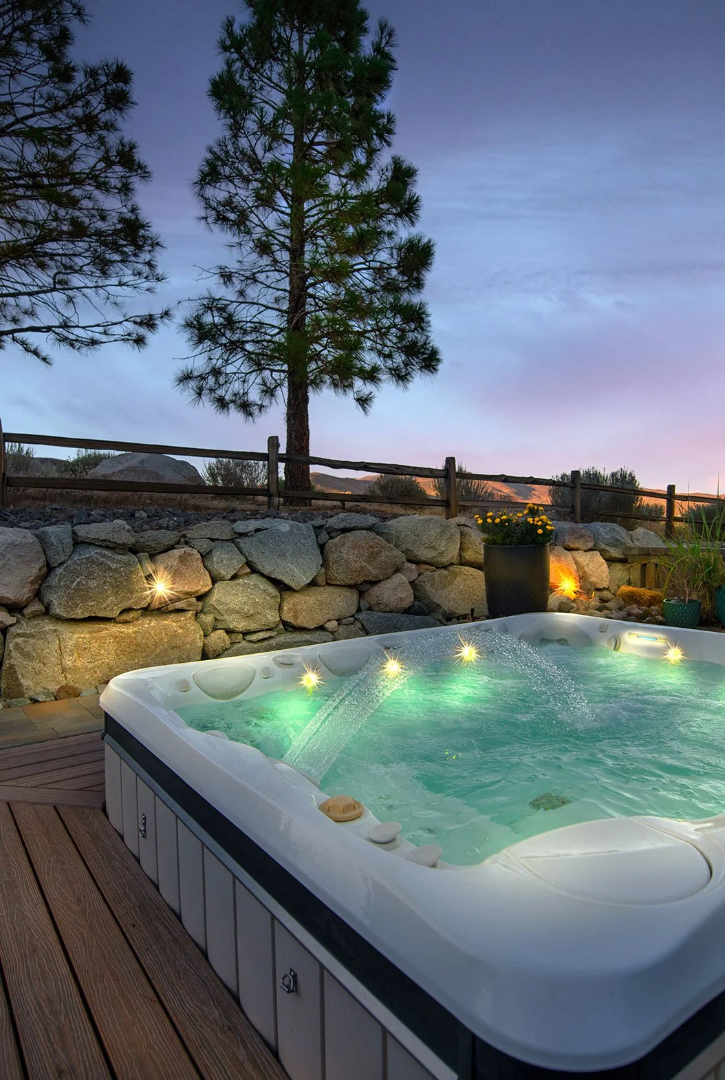 Outdoor hot tub with jets on a wooden deck, surrounded by a stone wall and potted plants, with a tall pine tree and a wooden fence in the background at dusk.