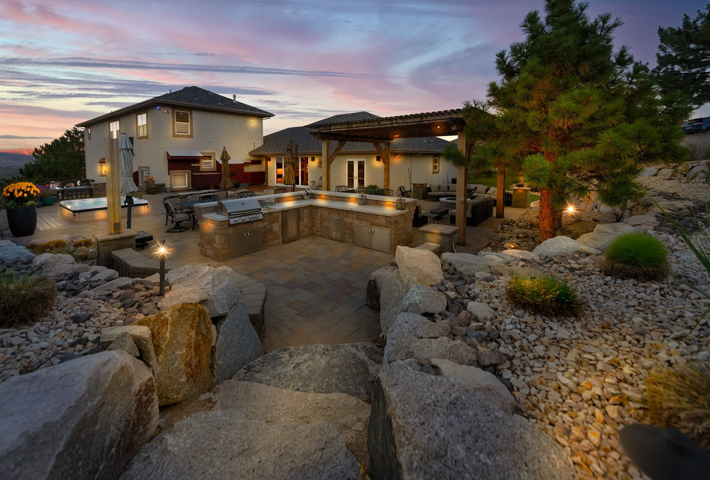 A spacious backyard patio at dusk featuring a built-in outdoor kitchen with a grill and counter space, a seating area with couches and umbrellas, a hot tub, a large tree, and landscaped rocks and plants, with the house and evening sky in the backgrou