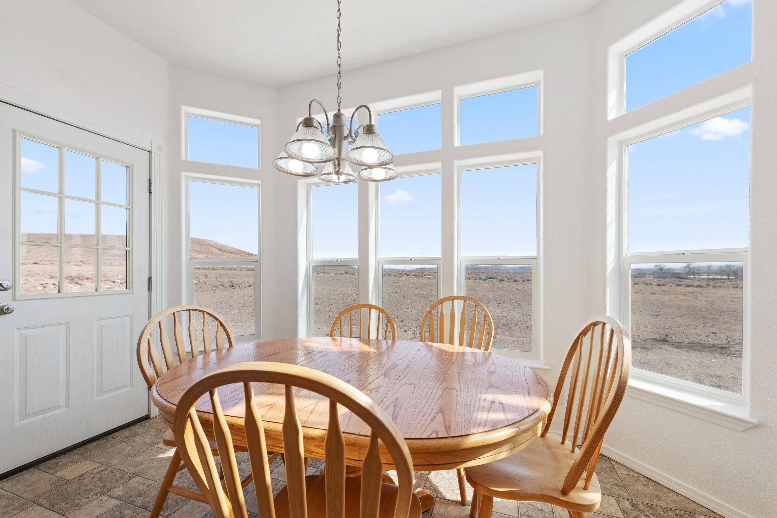 Kitchen with white walls and large windows, wooden dining table with six chairs, and a ceiling light fixture.