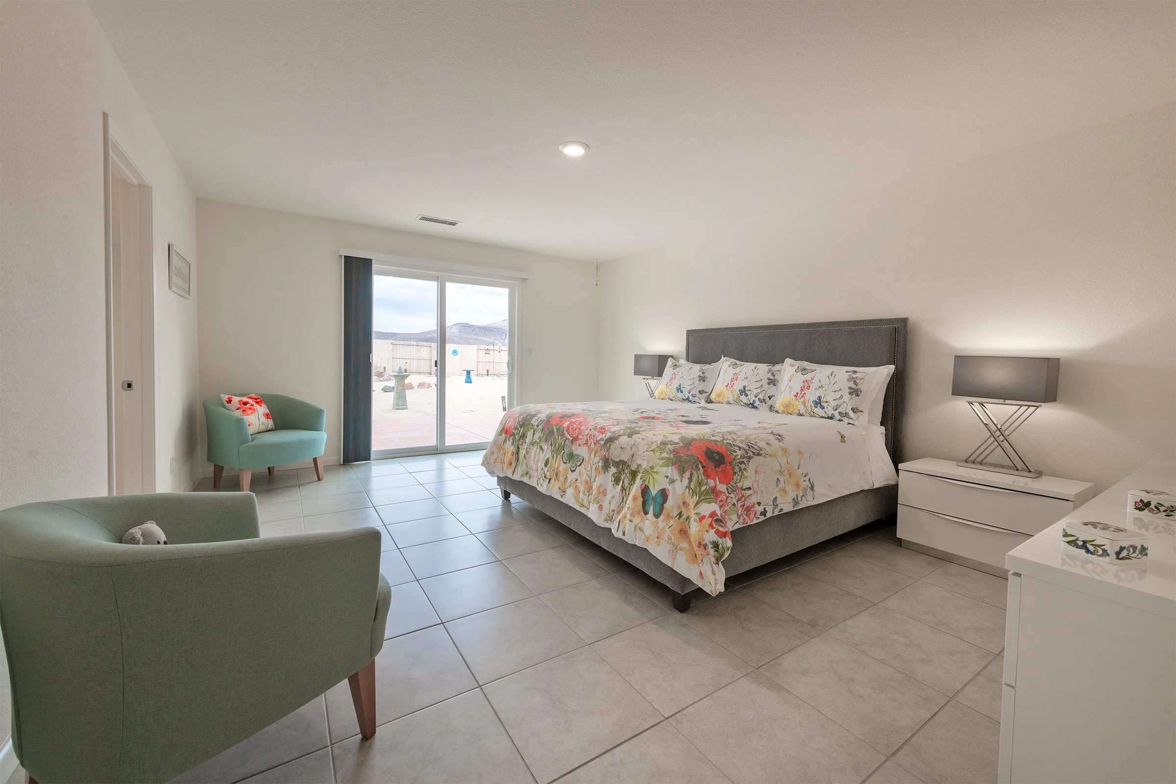 Bright bedroom with a large bed, floral bedding, two nightstands with lamps, two pastel-colored chairs, and a sliding glass door leading to an outdoor space with mountains in the background.