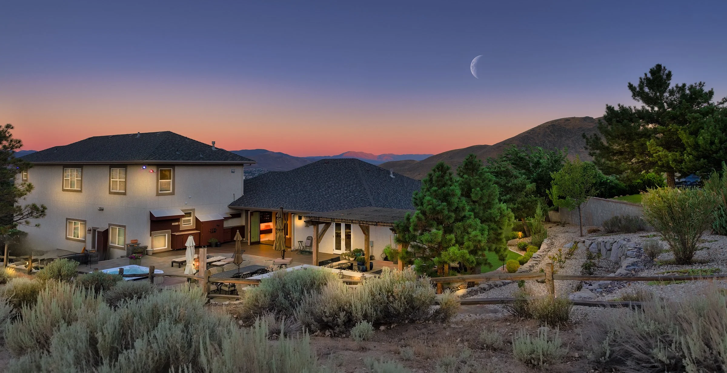 A sunset view of a backyard with outdoor furniture, a hot tub, trees, and a hillside, with the moon visible in the sky.