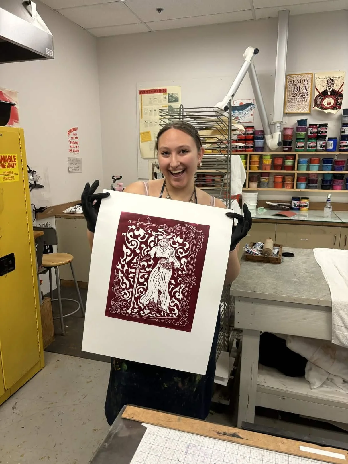 Young woman in a printmaking studio holding a red and white paper cut-out artwork of a woman with a staff, wearing a long flowing dress, surrounded by decorative patterns.