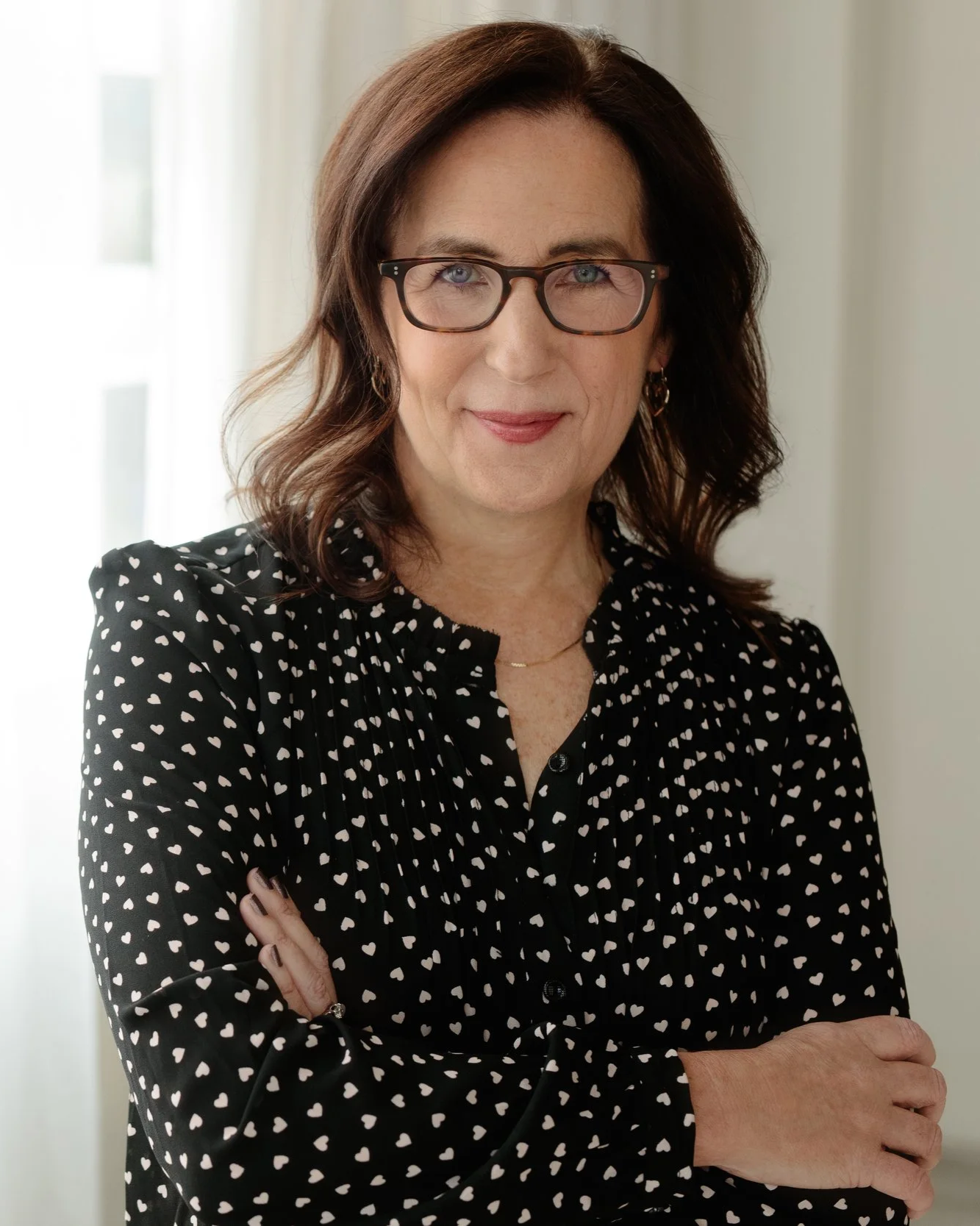 Leslie Wilson, founder and president of Lew Learning. A woman with dark brown hair and glasses, wearing a black blouse with white hearts, standing with her arms crossed in front of a light-colored background.