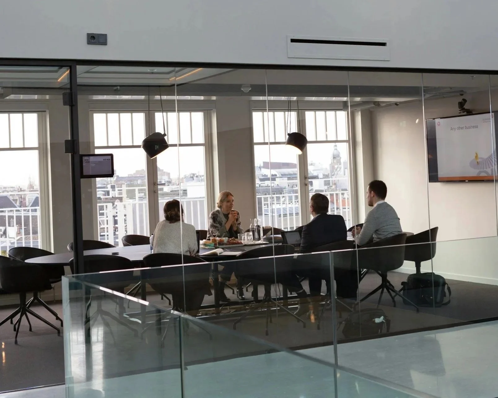 Business meeting with four people in a modern conference room, seen through glass partition, with cityscape outside windows.