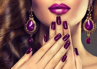 Close-up of a woman with purple lipstick and matching earrings, holding her hand near her lips showing purple manicured nails.