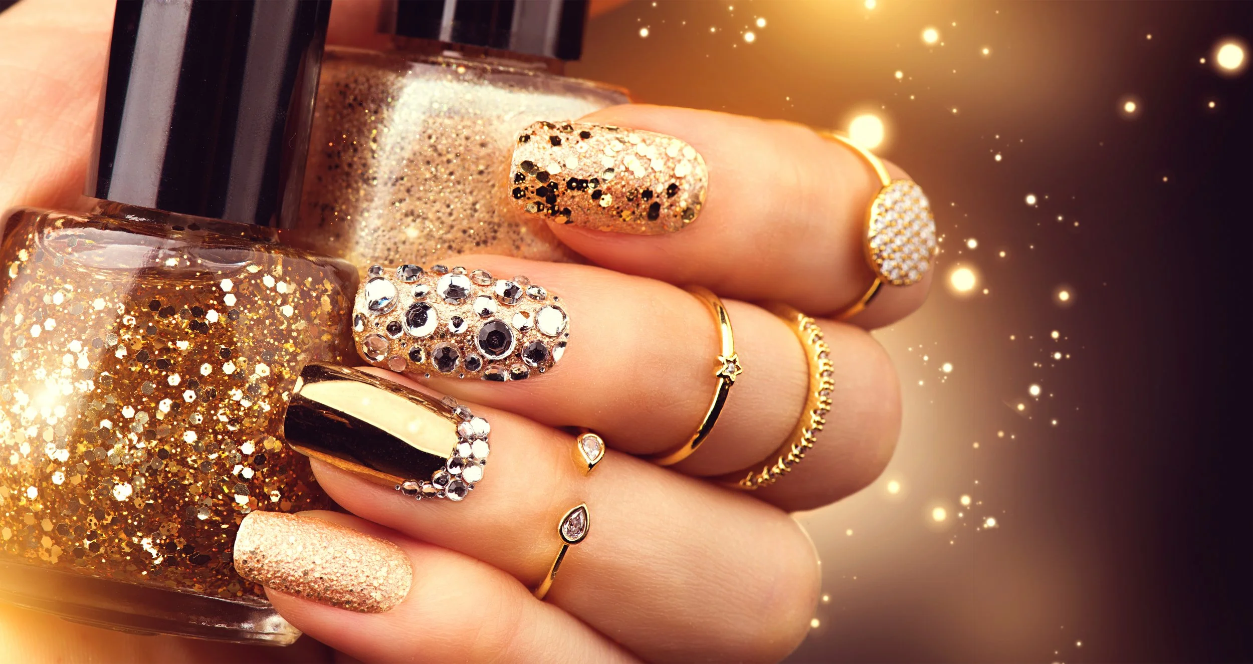 Close-up of a hand with decorated nails, wearing multiple gold rings, holding a bottle of gold glitter nail polish with gold and black glitter inside, with a background of glowing lights and sparkles.