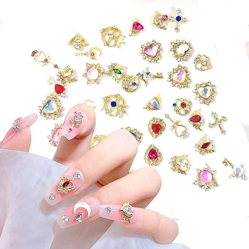 Multiple gold-colored charms with colorful gemstones and decorative designs scattered on a white surface, with a hand showing off decorated nails wearing matching charms.